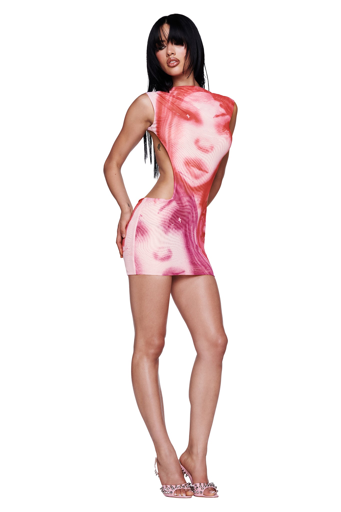 A woman with long black hair wears the I.AM.GIA T-REX CORALINE MINI DRESS in pink, a sleeveless micro mini dress with face prints that’s perfect for festival season. She poses in high heels with one hand on her hip against a white background.