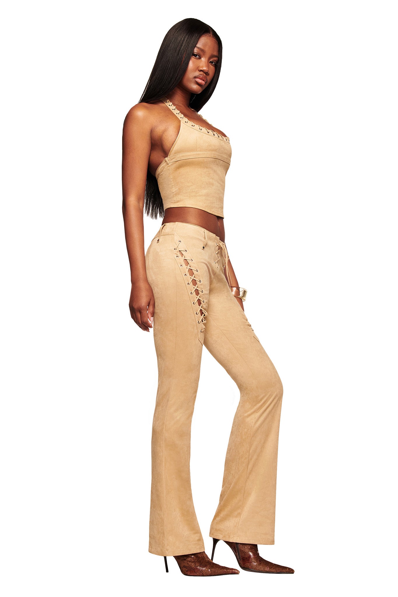 A woman in profile wears the I.AM.GIA XENIA SUEDE PANT - TAN with a matching crop top, long straight hair, and pointed brown heeled boots, set against a white background.