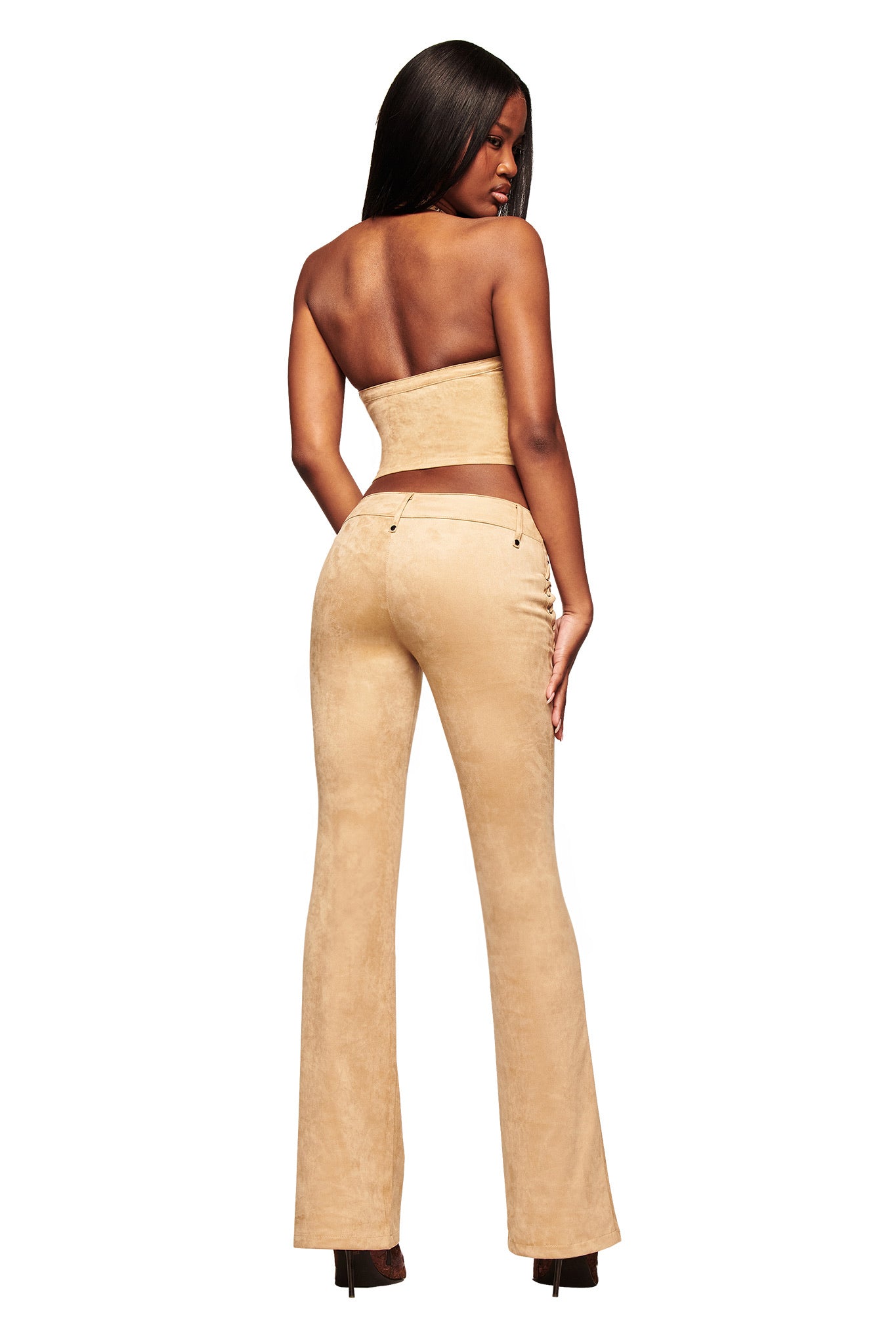 A woman with long dark hair stands with her back to the camera, wearing a strapless beige crop top and I.AM.GIA XENIA SUEDE PANT in tan with heels. The background is white.
