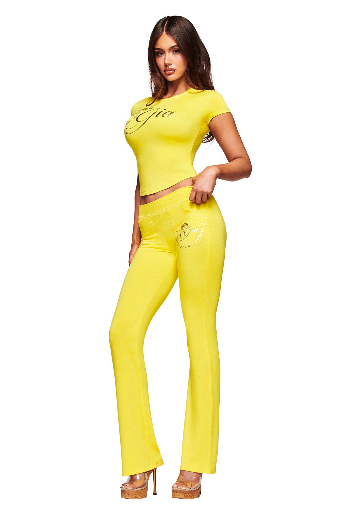 A woman poses in I.AM.GIA’s BLARE FORM POCKET TRACKPANT in yellow gold foil, featuring flared legs and script details. She pairs it with a matching short-sleeve crop top, high heels, and her long dark hair styled in loose waves.