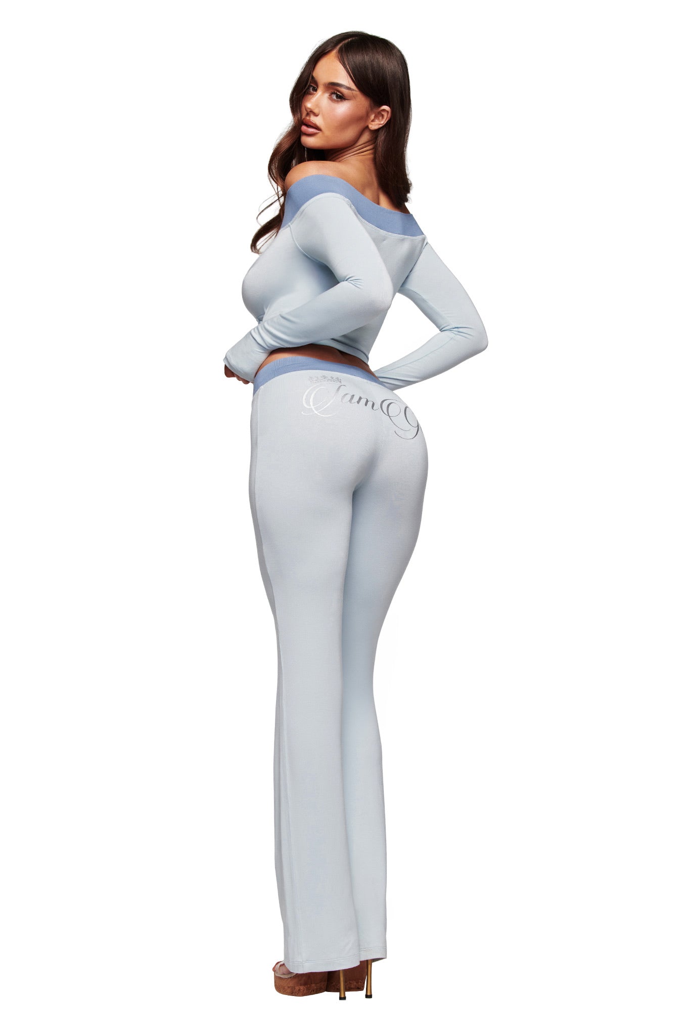 A woman with long brown hair models I.AM.GIA's BLARE LOWER RISE TRACKPANT in two tone powder blue, pairing the sporty, flared-leg pants with a matching fitted top and high heels, looking over her shoulder against a white background.