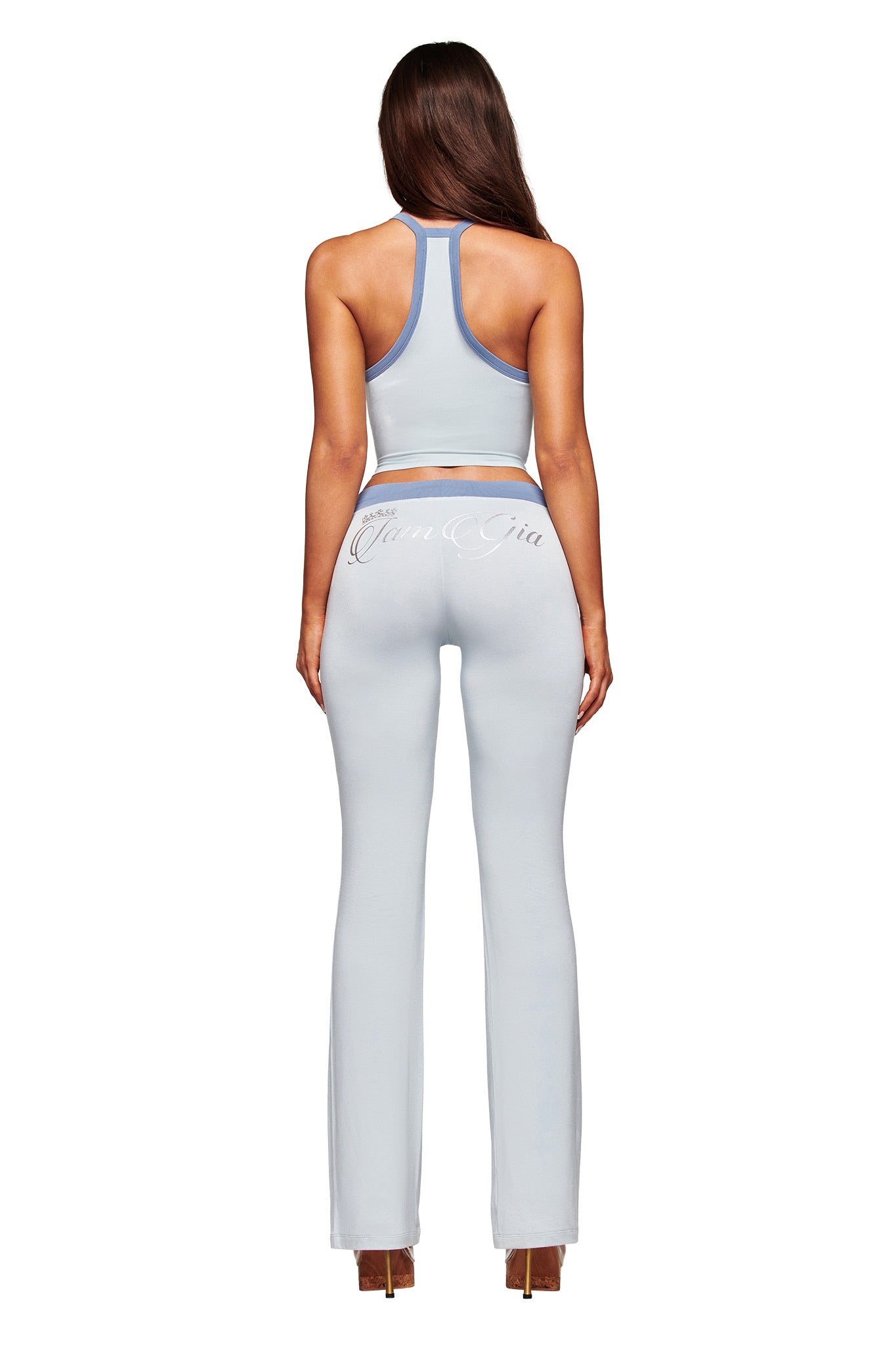A woman faces away wearing I.AM.GIA's light blue BLARE LOWER RISE TRACKPANT - TWO TONE POWDER BLUE with flared legs and script waistband, paired with a matching sleeveless crop top featuring a racerback design and heels.