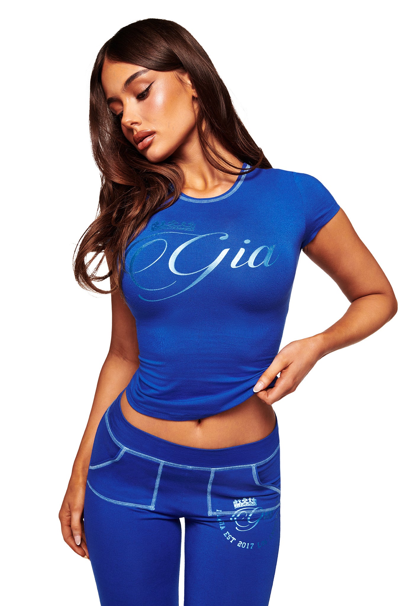 A woman with long brown hair poses against a white background, wearing the BLARE FORM TEE - COBALT FOIL from I.AM.GIA—a body-hugging cobalt blue bamboo tee with “Gia,” paired with matching faux pocket leggings.
