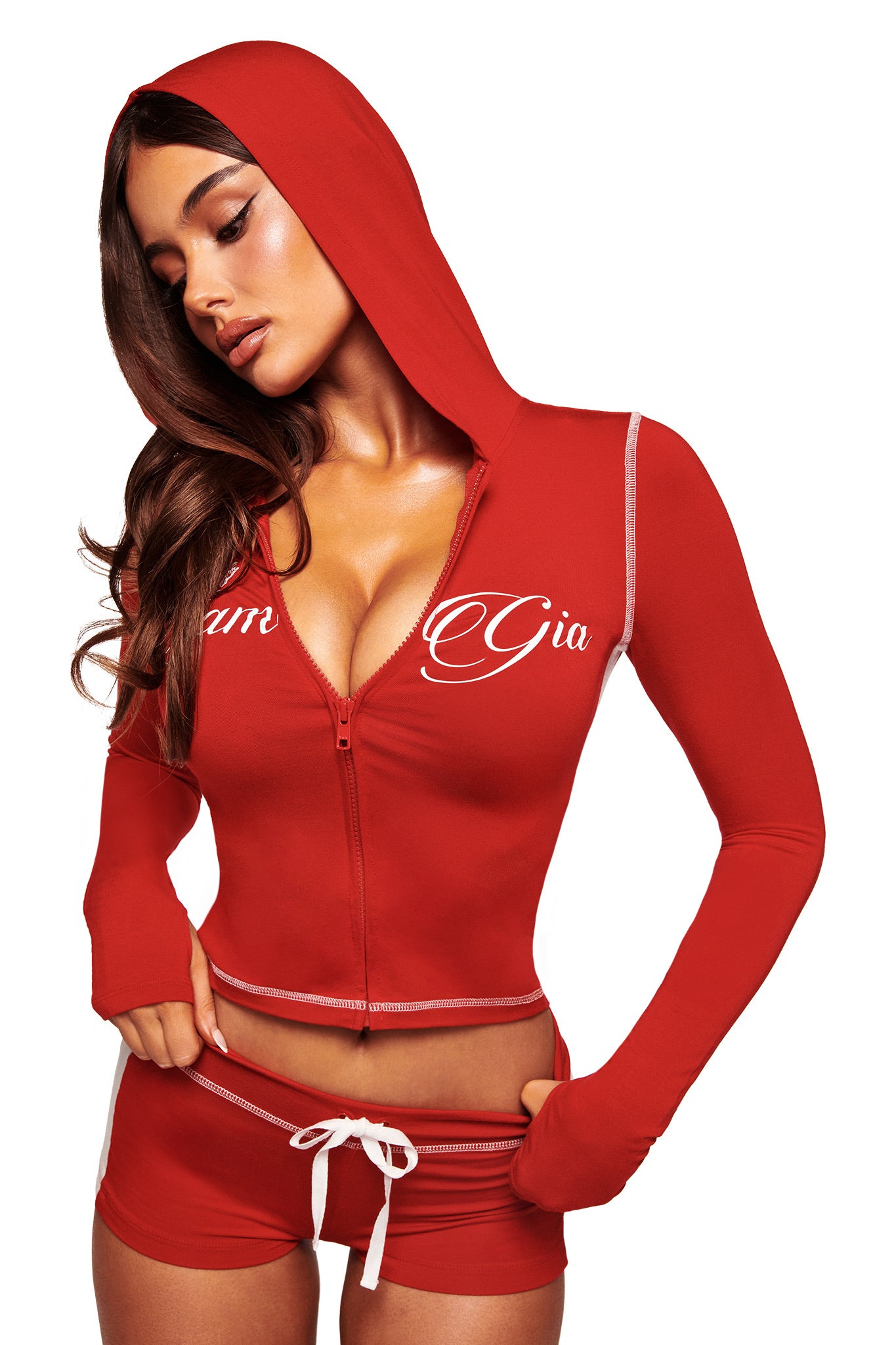 A woman poses in a red I.AM.GIA hooded zip-up crop top and the BLARE PANELLED SHORT - RED, featuring sporty white trim and a drawstring. "Gia" and "amr" in white script appear on the top as she looks down, one hand resting on her shorts.