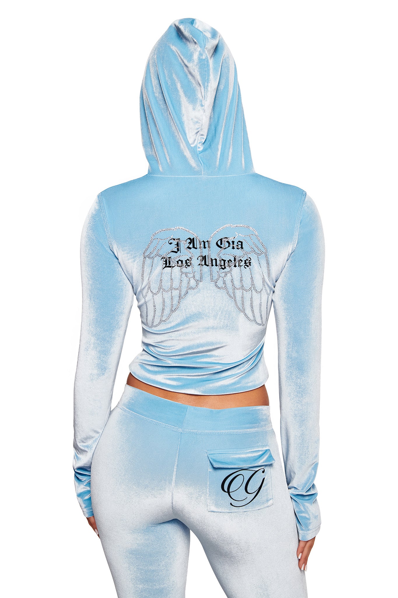 Wearing I.AM.GIA’s BLARE VELOUR POCKET TRACKPANT in ice blue, featuring a small decorative "G" pocket, plus a matching light blue velour hooded jacket with angel wings and “I Am Gia Los Angeles” on the back.