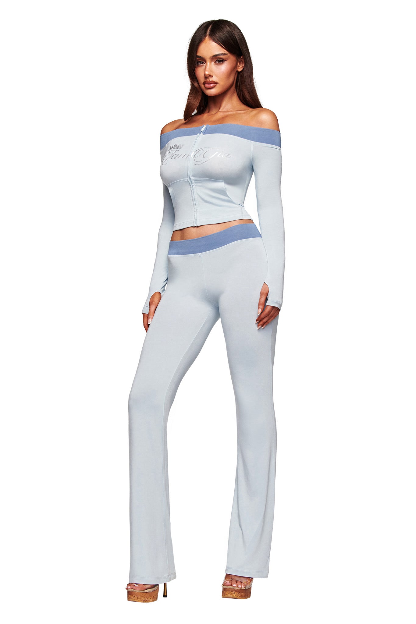 A woman poses in I.AM.GIA’s BLARE LOWER RISE TRACKPANT - TWO TONE POWDER BLUE with blue trim, a matching off-the-shoulder crop top, and clear heels against a white background.