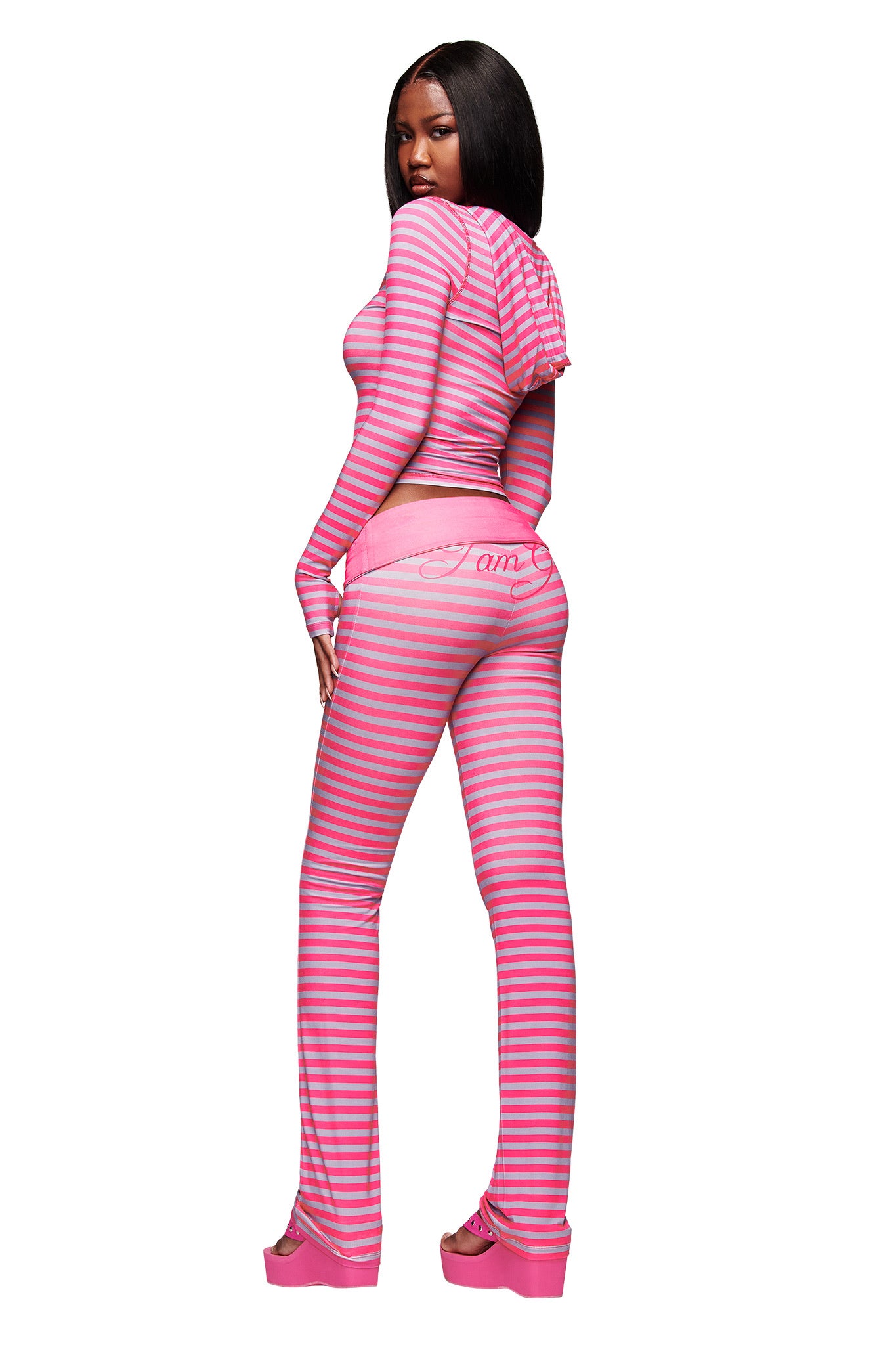 A woman faces backward in I.AM.GIA’s BLARE FOLDOVER TRACKPANT - HOT PINK STRIPE, a fitted, long-sleeve set with flared legs. She wears platform shoes and has straight, long dark hair against a white background.