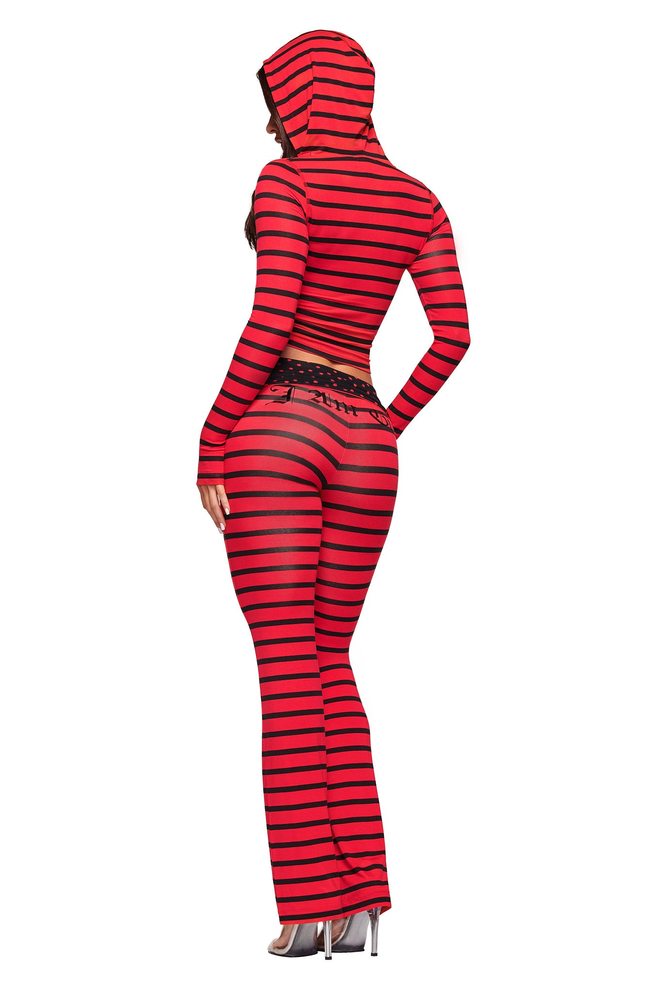 A person wearing I.AM.GIA’s BLARE FOLDOVER TRACKPANT in red and black stripes, paired with a matching sheer hooded top, stands with their back to the camera and wears high-heeled shoes.
