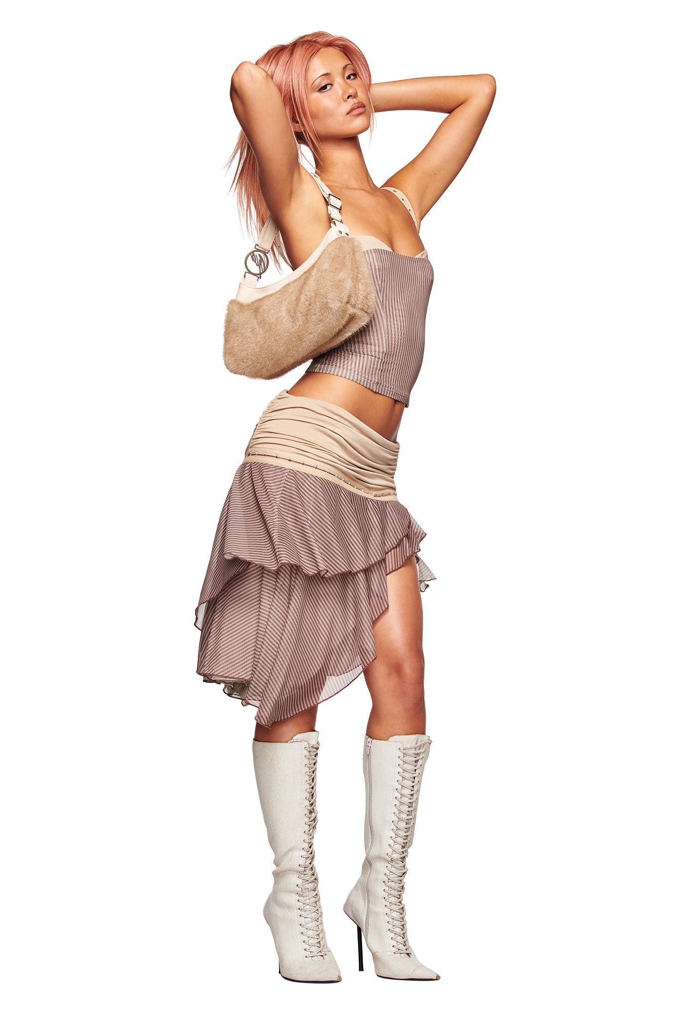 A woman with pink hair poses with arms raised, wearing the I.AM.GIA SEPHI MIDI SKIRT in Mushroom Stripe, white heeled lace-up boots, and a beige faux-fur shoulder bag against a white background.