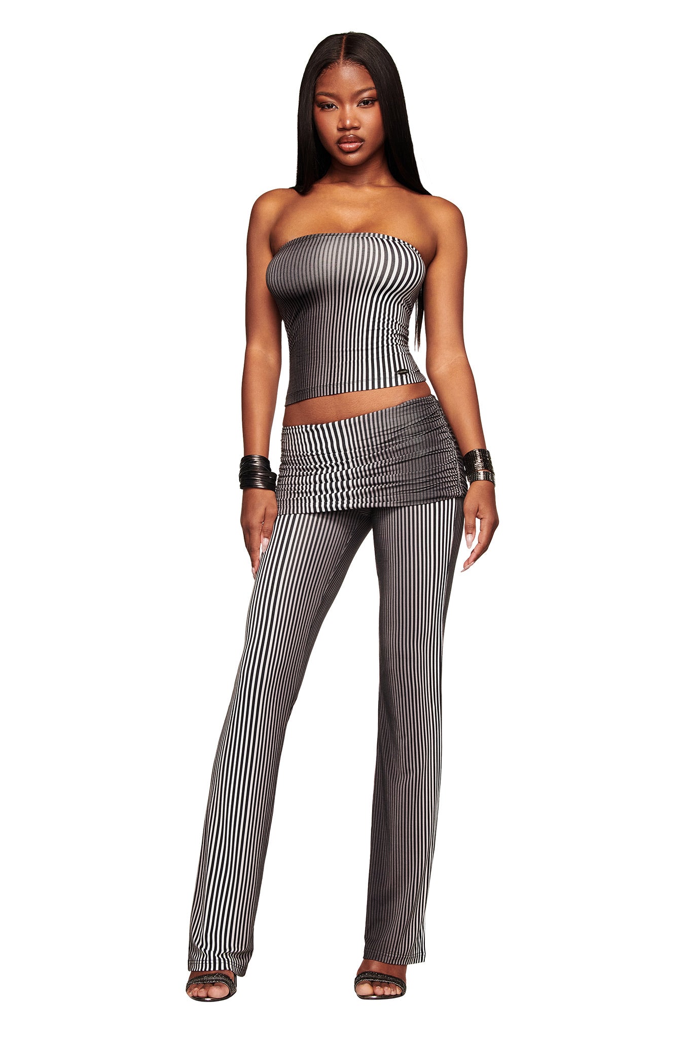 A woman faces forward in the I.AM.GIA TRINITY 2-IN-1 SKIRT PANT - STRIPE, featuring a black and white ombre stripe print strapless top, with long straight hair, black wristbands, open-toe heels, and a white background.