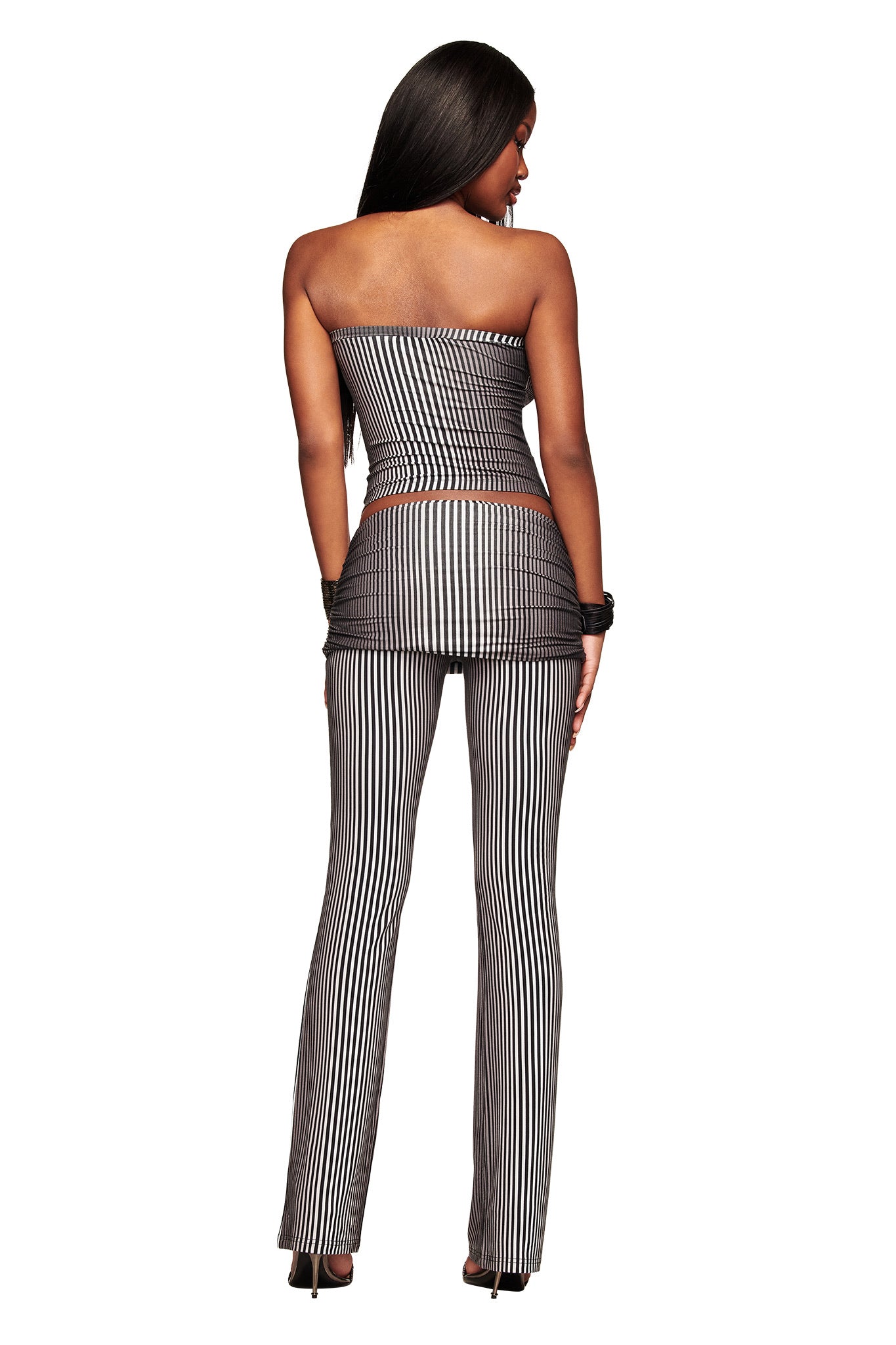 A woman faces away, wearing the I.AM.GIA TRINITY BOOB TUBE - STRIPE in a black and white ombre stripe bodycon top and matching flared pants with black heels, set against a white background.