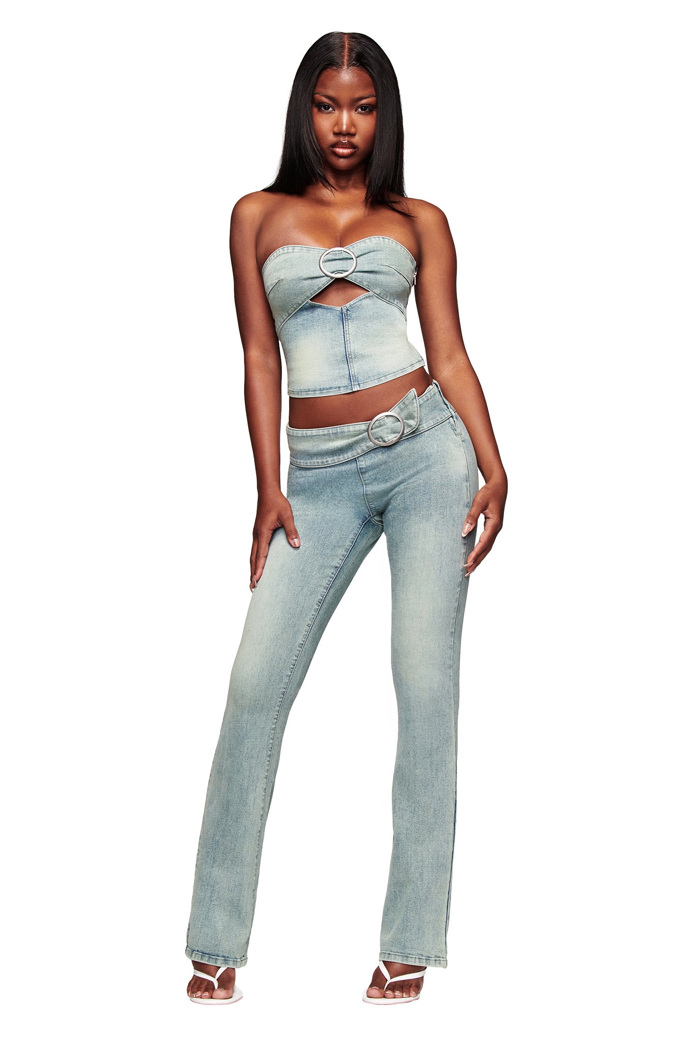 A woman stands confidently in I.AM.GIA's JENN JEAN - BLUE, featuring a strapless light denim top with a heart cutout and matching light wash, mid-rise flared jeans, paired with clear high-heeled sandals.