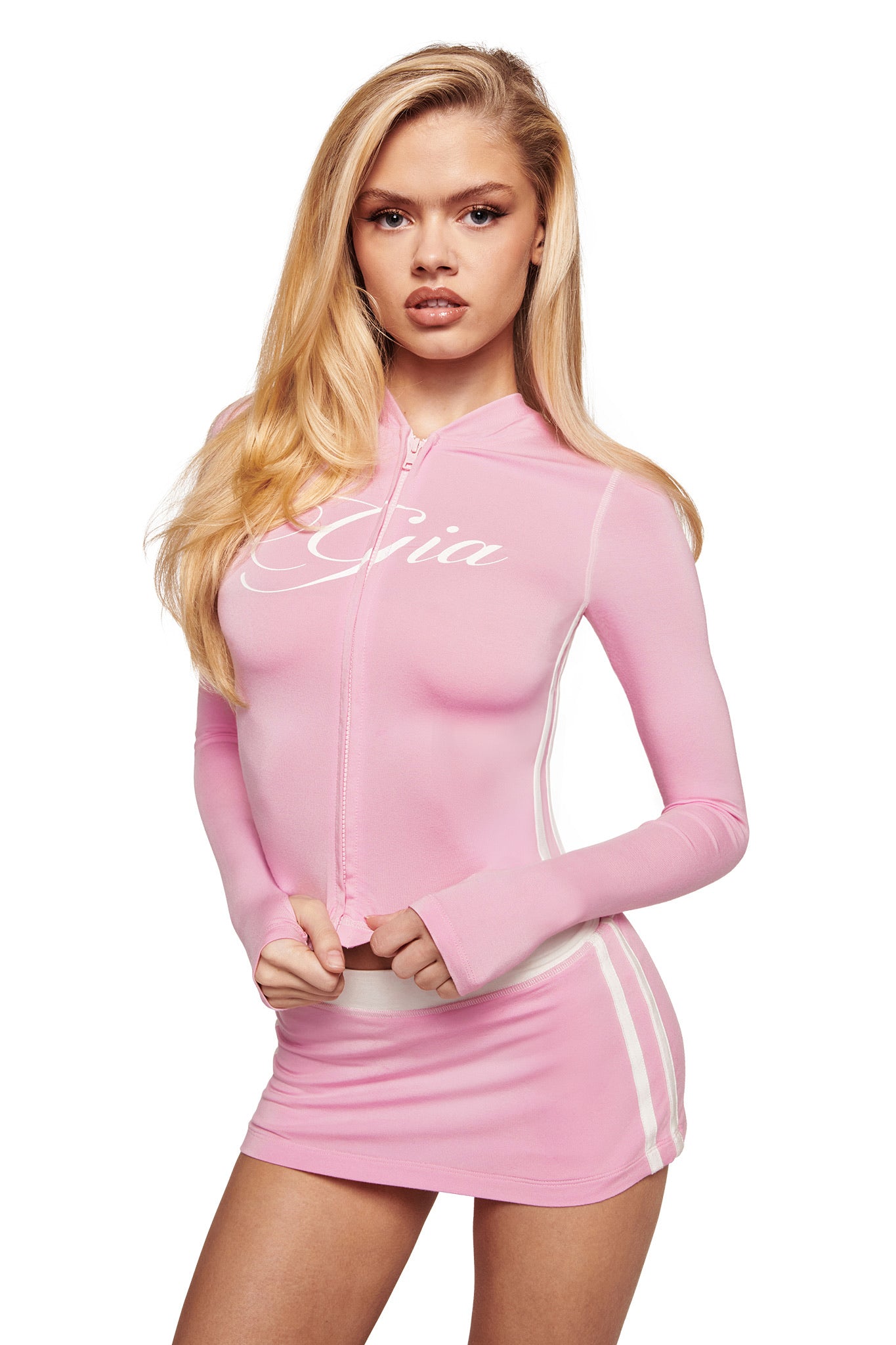 A woman with long blonde hair wears a light pink zip-up hoodie with "Gia" and the I.AM.GIA BLARE PIPING MICRO SKORT in baby pink and white, featuring white stripes, standing against a white background.