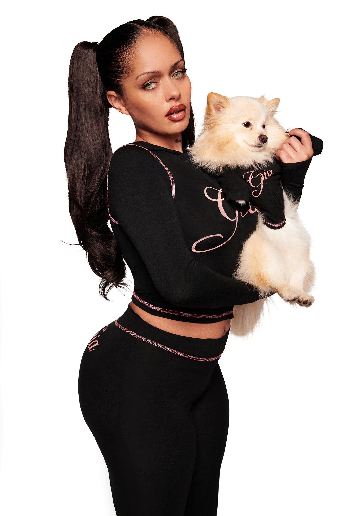 A woman with long dark hair in pigtails wears a fitted black outfit with pink trim, holding a fluffy white dog dressed in the I.AM.GIA BLARE BEST FRIEND DOG HOODIE - BLACK. Both pose against a plain white background.