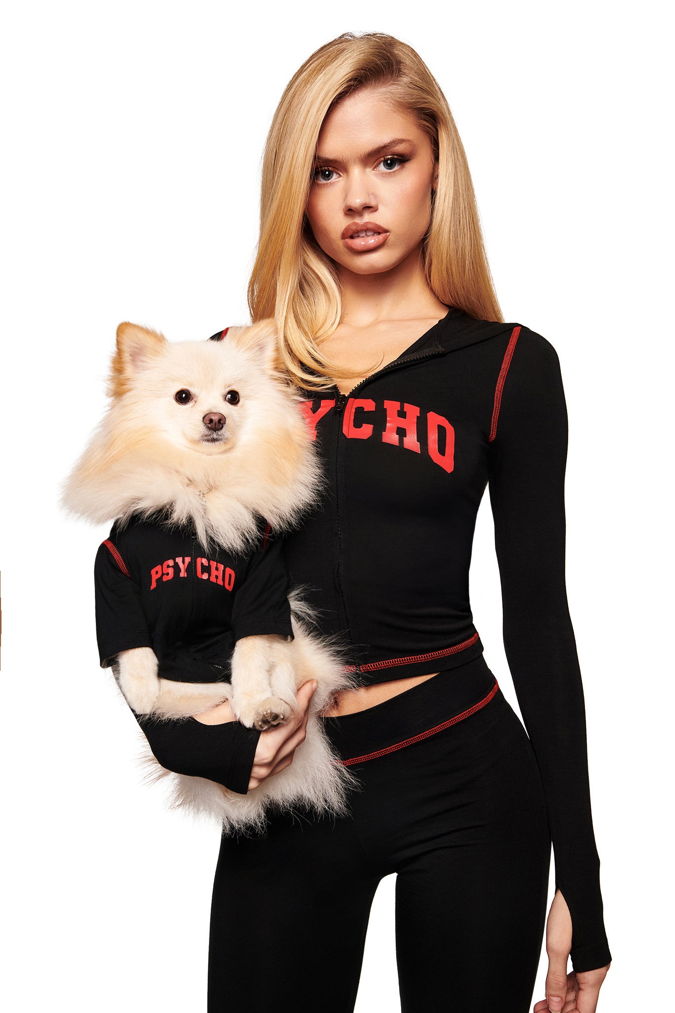 A woman wears the I.AM.GIA BLARE BEST FRIEND DOG HOODIE in black with "PSYCHO" print, holding a fluffy white dog in a matching hoodie. Both face forward against a white background.