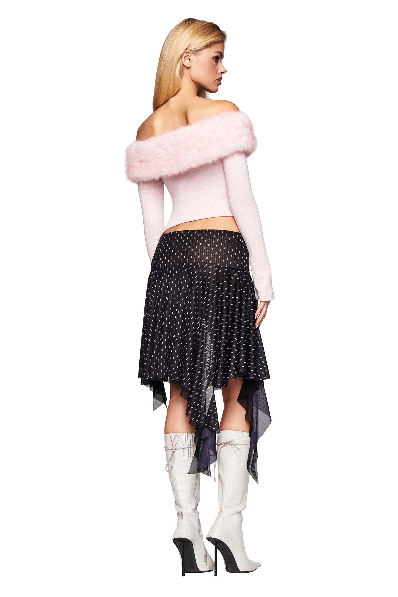 A person faces away in the I.AM.GIA CECILY TOP - PINK, a ribbed off-shoulder long sleeve with furry trim, paired with a black asymmetrical polka dot skirt and white knee-high heeled boots. The background is plain white.