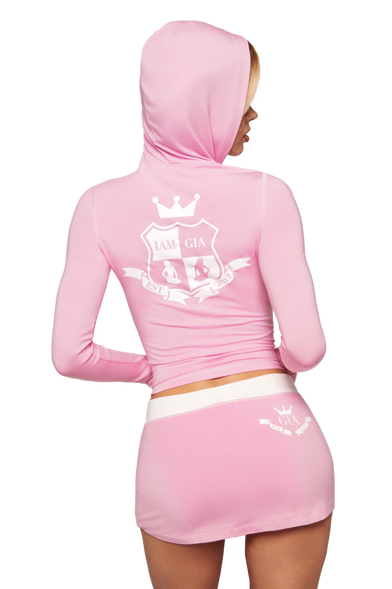 A person wearing the I.AM.GIA BLARE PIPING MICRO SKORT in baby pink and white, paired with a matching hooded crop top featuring white printed crest designs, stands facing away from the camera.