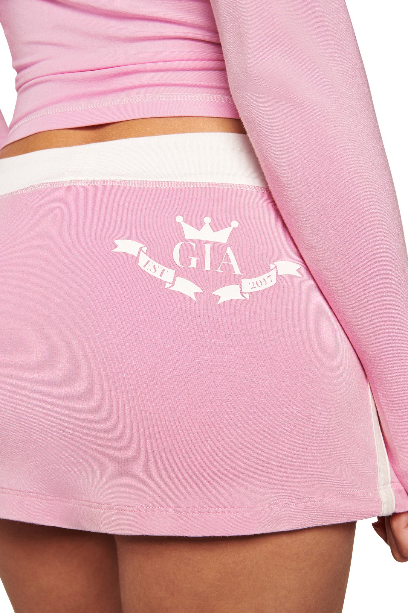 A person wears the I.AM.GIA BLARE PIPING MICRO SKORT in baby pink and white with a sporty design, featuring a white stripe and back graphic of a crown, "GIA," and banners reading "Est." and "2017", paired with a matching top.