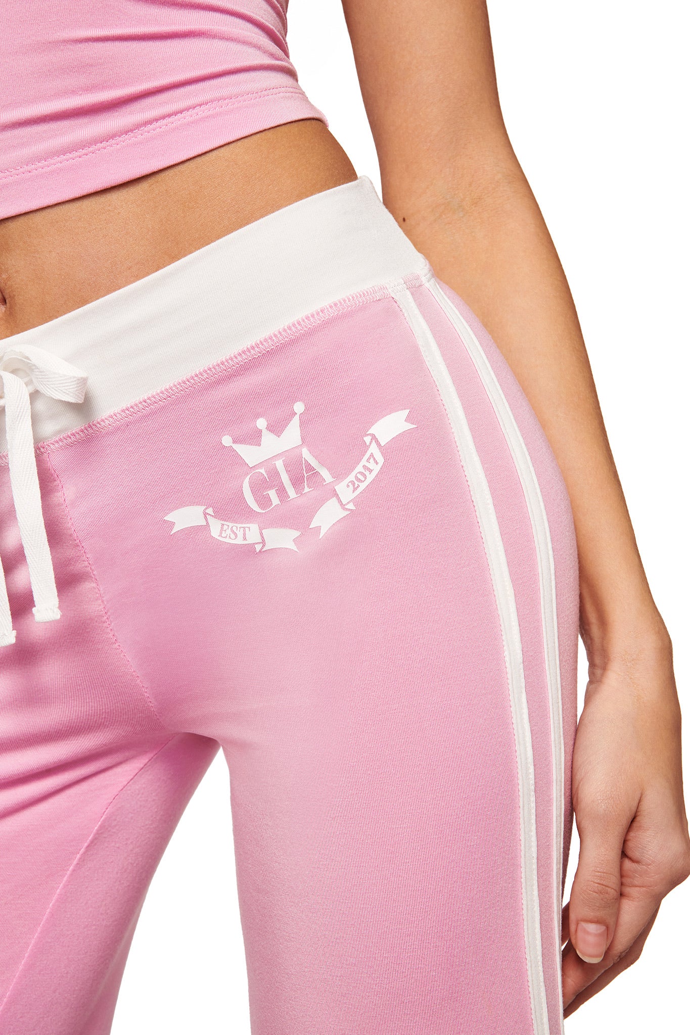 A person wears I.AM.GIA's BLARE PIPING TRACKPANT in baby pink and white, featuring white stripes, a white waistband, straight legs, a crown graphic, and "GIA EST 2017" on the hip. Their arm is visible.