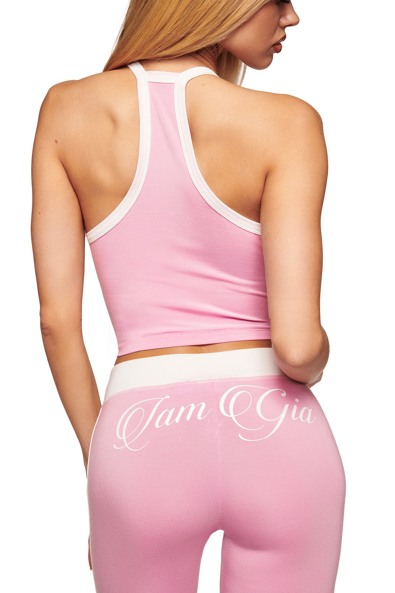 A woman with long blonde hair faces away, wearing the I.AM.GIA BLARE V-NECK TANK in baby pink and white racerback style, paired with matching high-waisted leggings. "I Am Gia" is written in white script on the back.