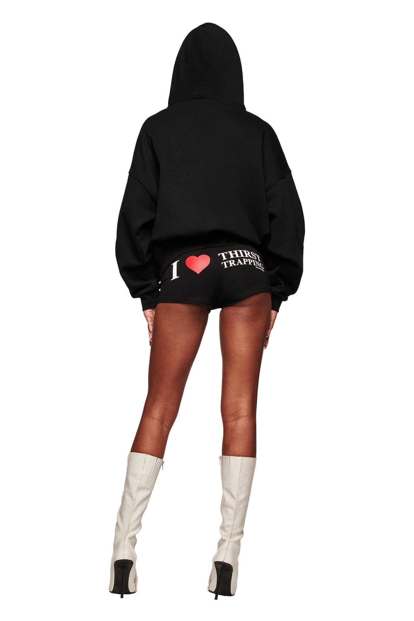 A person wears the I.AM.GIA POLINA HOODIE - THIRST TRAPPER, black shorts with white and red text, and white knee-high boots, standing with their back to the camera against a white background.