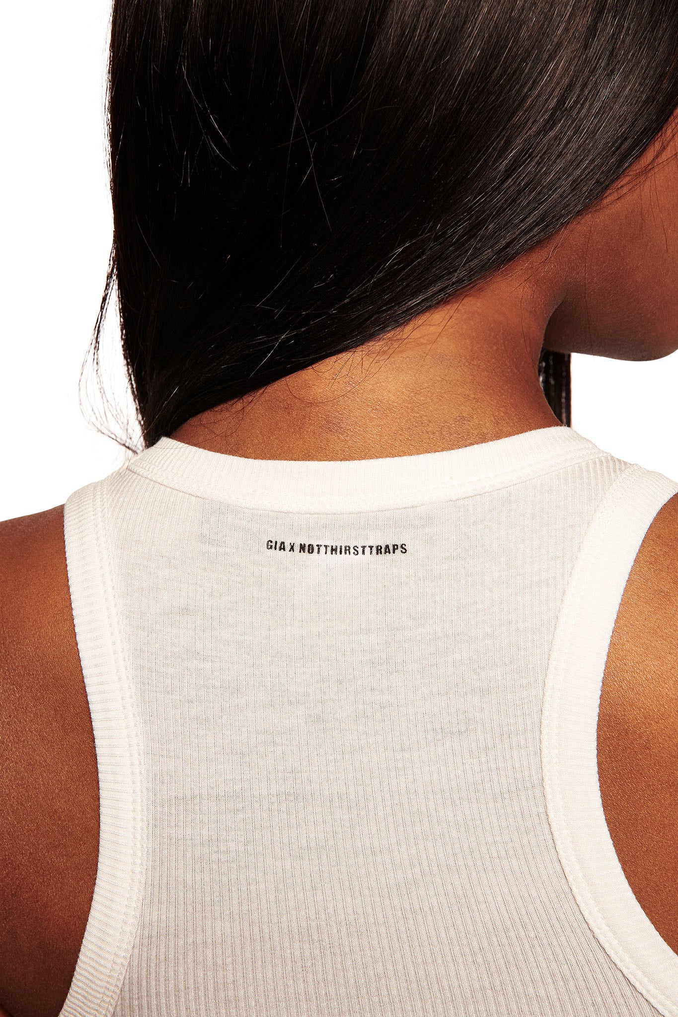 A close-up of a person in the I.AM.GIA POLINA TANK - MANIPULATOR, highlighting the white rib knit back neckline with “GIA X N0THESTRAPS” in small black letters and partially visible long, dark hair.