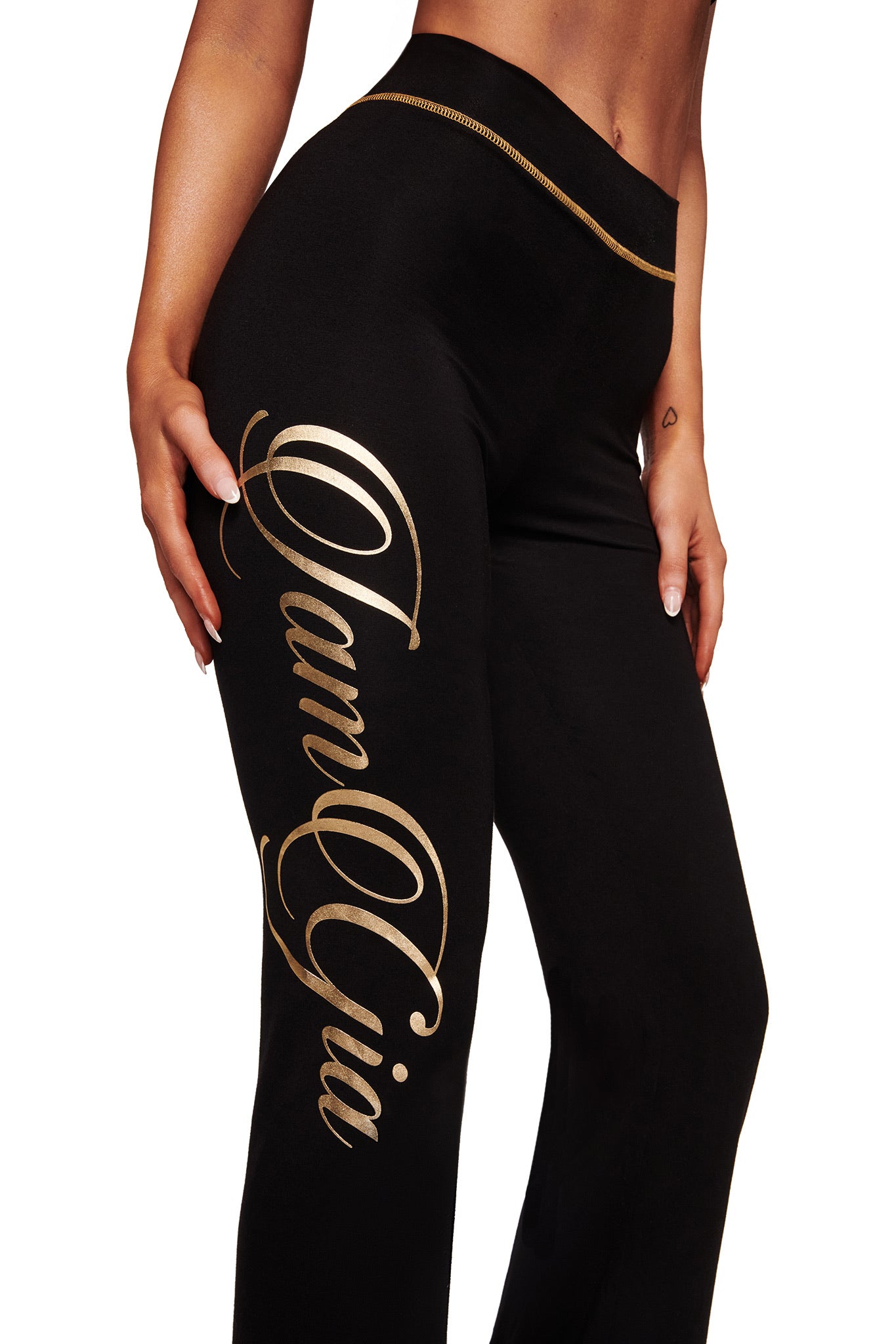 A woman wears the I.AM.GIA BLARE FORM TRACKPANT in black and gold, featuring bold gold "I.AM.GIA" script down one leg. The sculpted Form fabric hugs her figure, highlighting her silhouette. Only her torso and legs are shown.