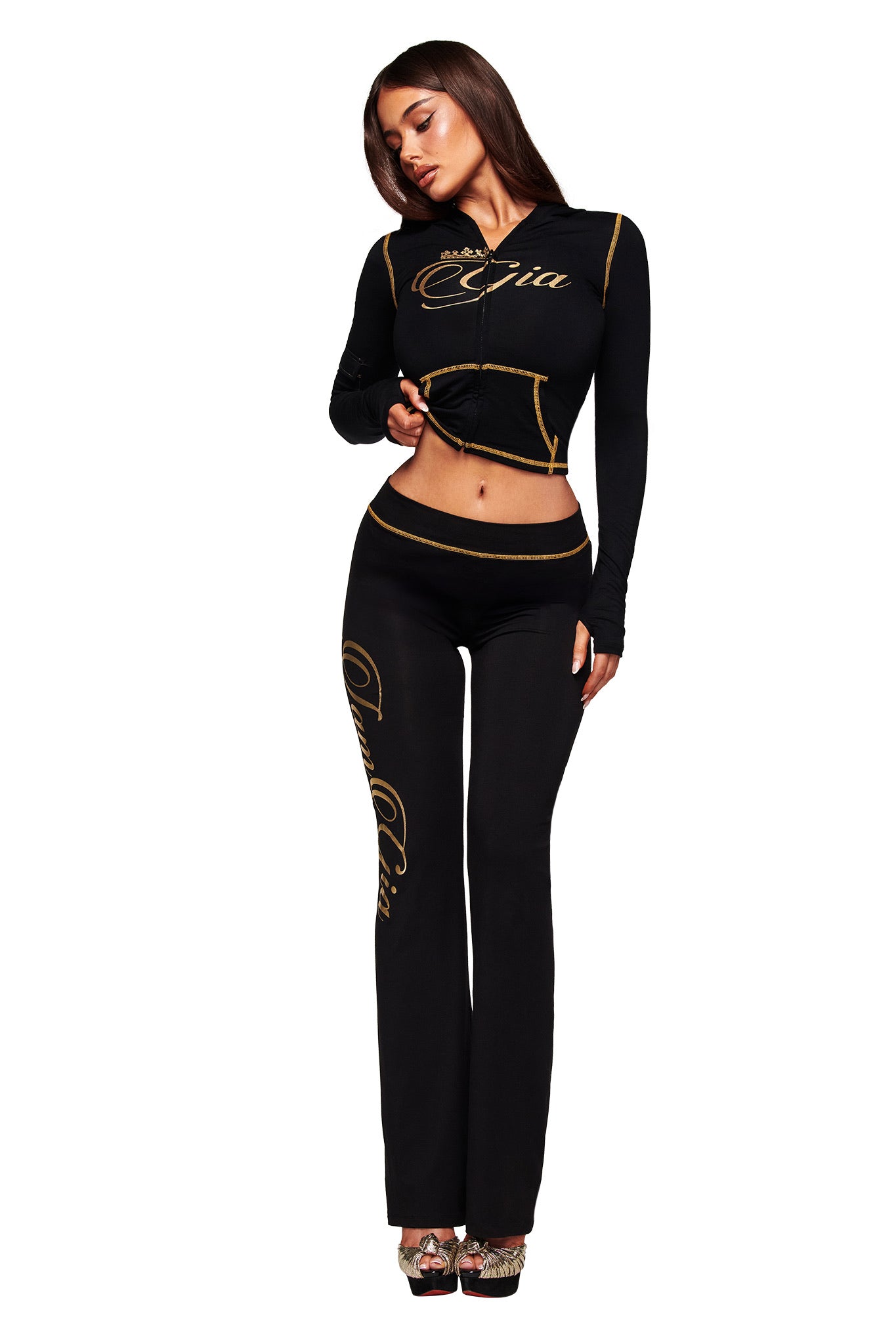 A woman poses against a white background in the I.AM.GIA BLARE FORM MULTI POCKET HOODIE in black and gold, featuring cursive gold lettering. She pairs it with high-waisted pants and high-heeled shoes.