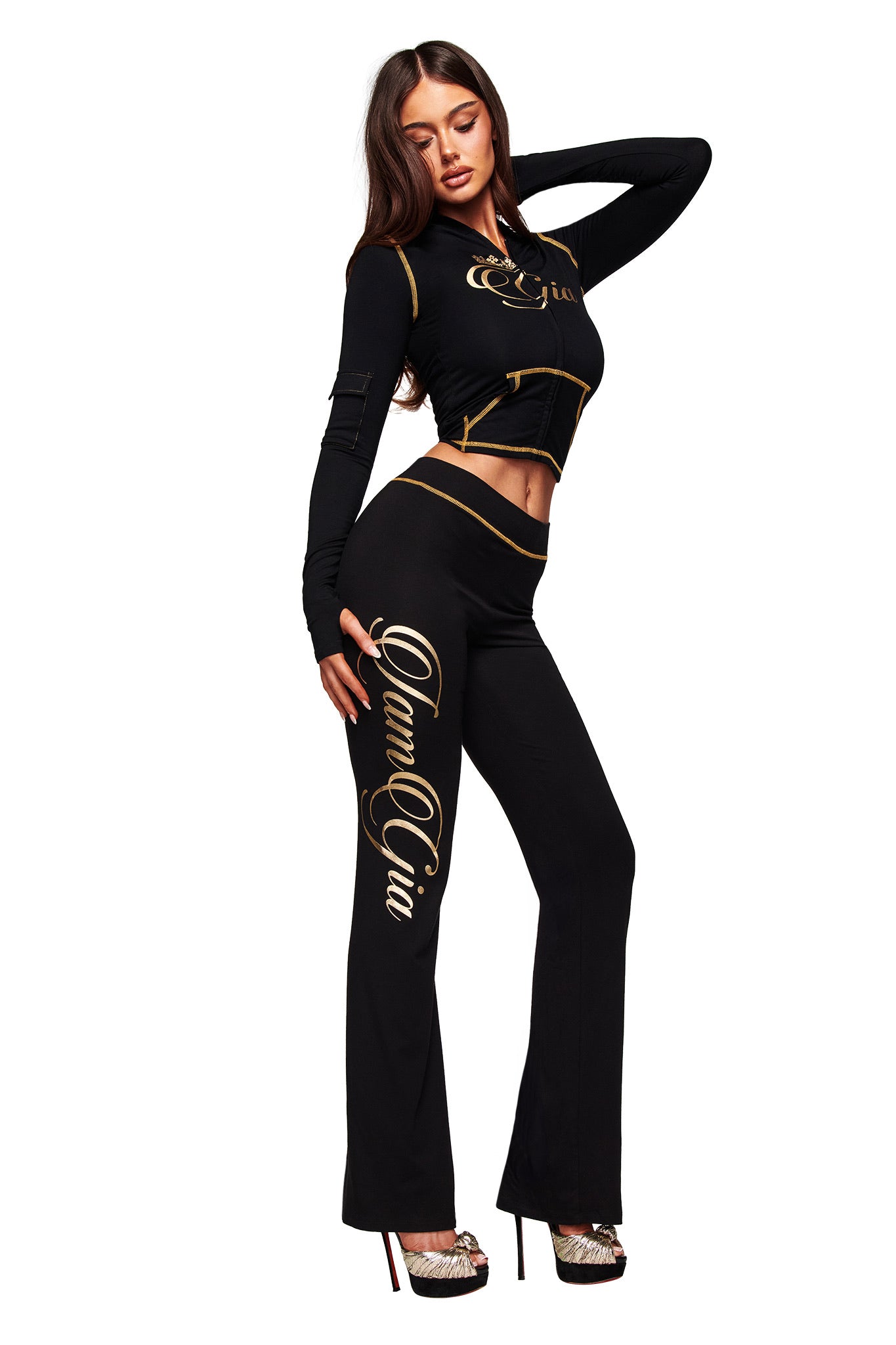 A woman models the I.AM.GIA BLARE FORM MULTI POCKET HOODIE in black and gold. She poses stylishly with high heels and long, dark hair, one hand on her head and the other on her hip.