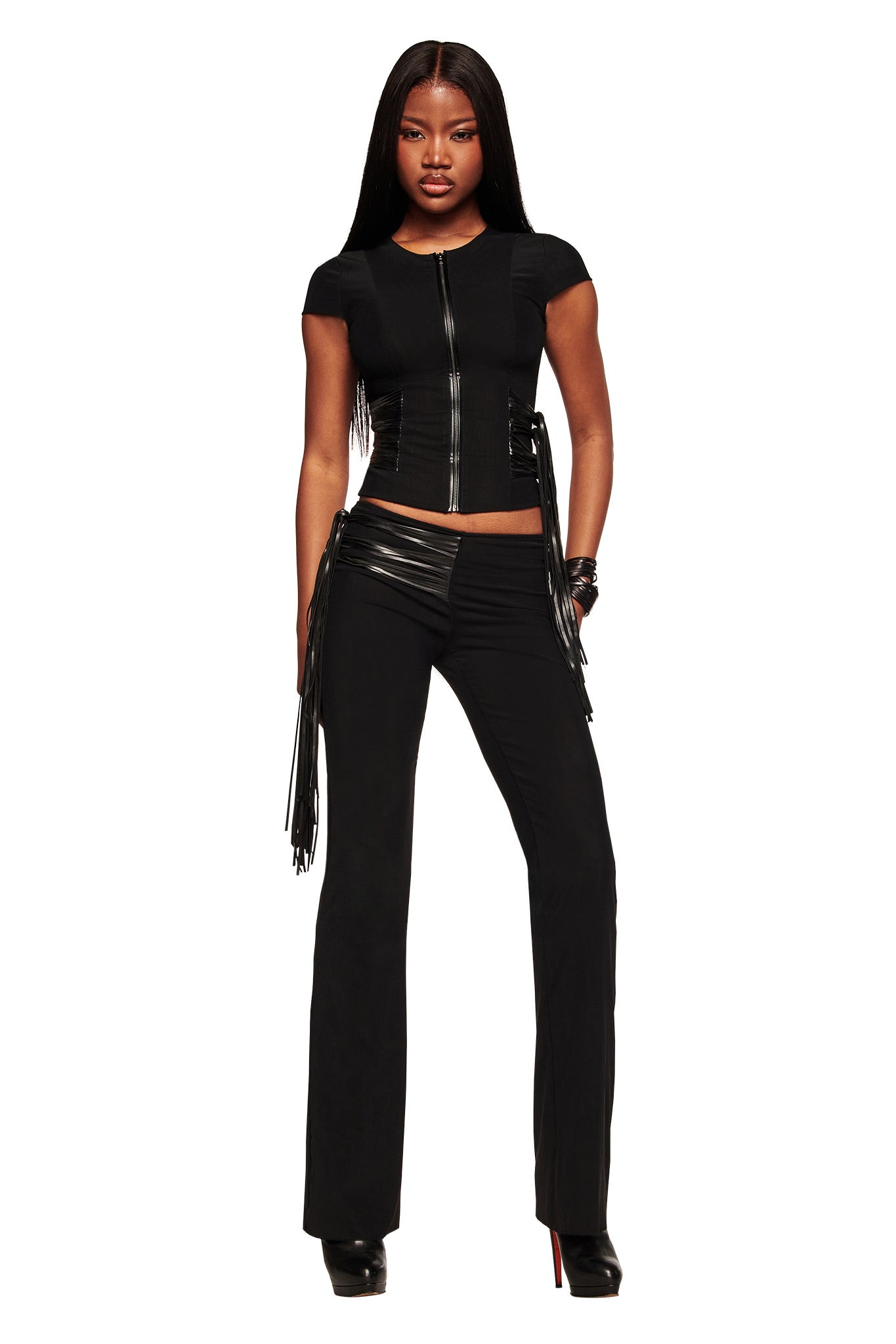 A woman exudes confidence in the I.AM.GIA ISELDA FRINGE TOP - BLACK, styled with high-waisted pants with faux leather fringe, black platform heels, and long, straight hair.