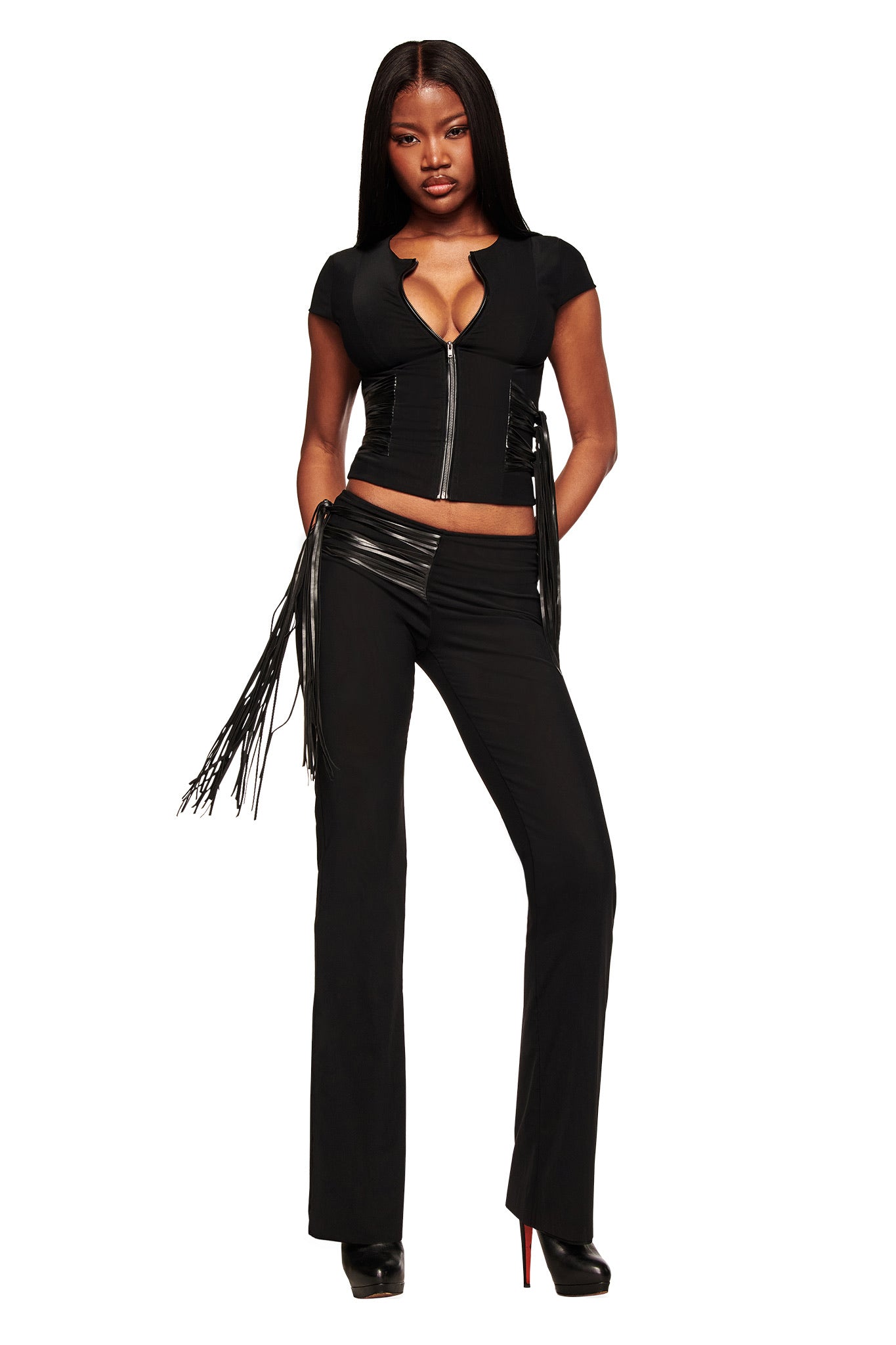 A woman with long straight hair models the I.AM.GIA ISELDA FRINGE PANT in black, featuring high-waisted shiny faux leather fringe, paired with a black short-sleeve zip-up crop top and black heels against a white background.