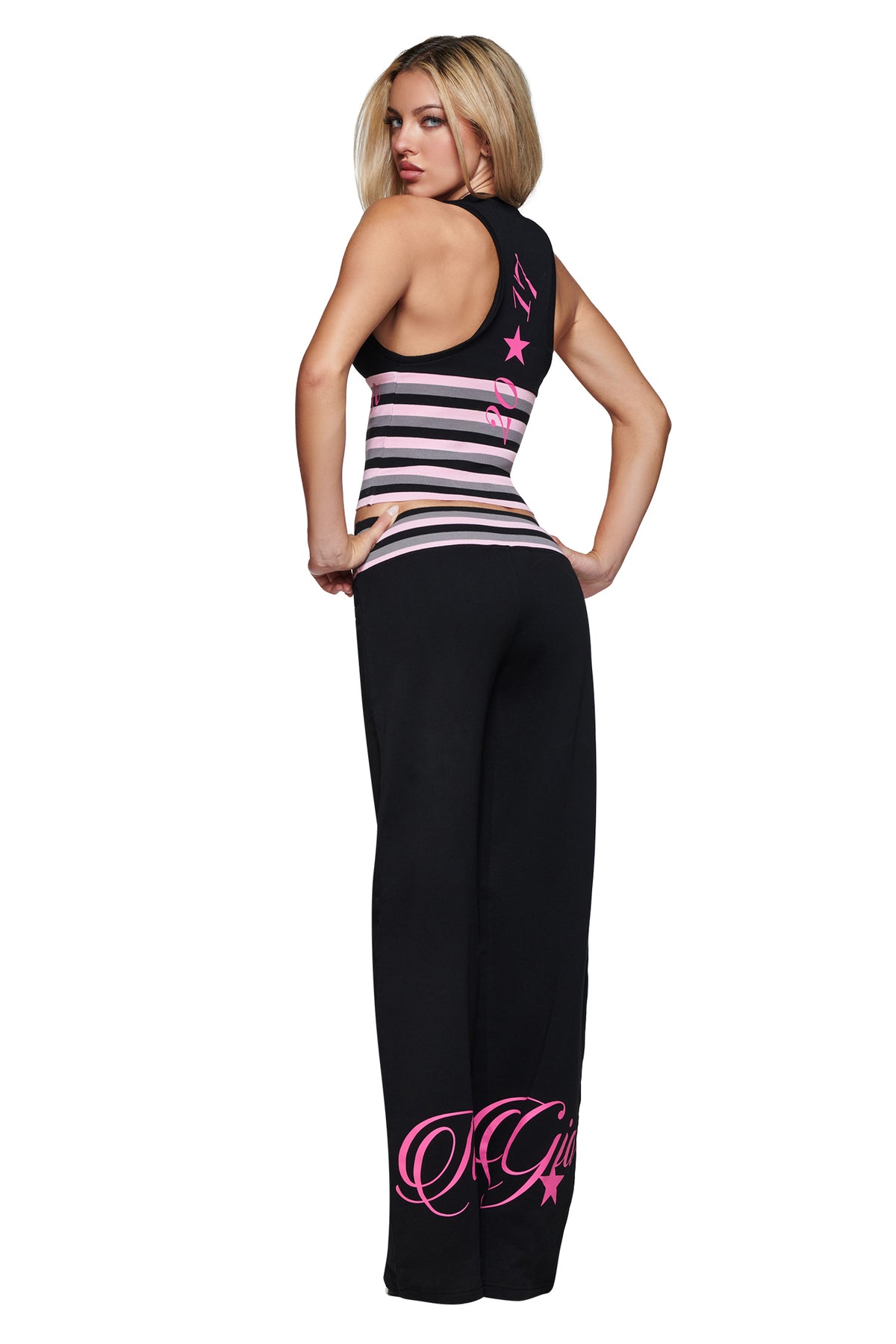 A woman with long blonde hair stands against a white background, looking over her shoulder while wearing the I.AM.GIA CANDI PANT in black—featuring pink writing, a comfortable mid-rise waist, and wide legs—paired with a black sleeveless top with pink and white stripes.