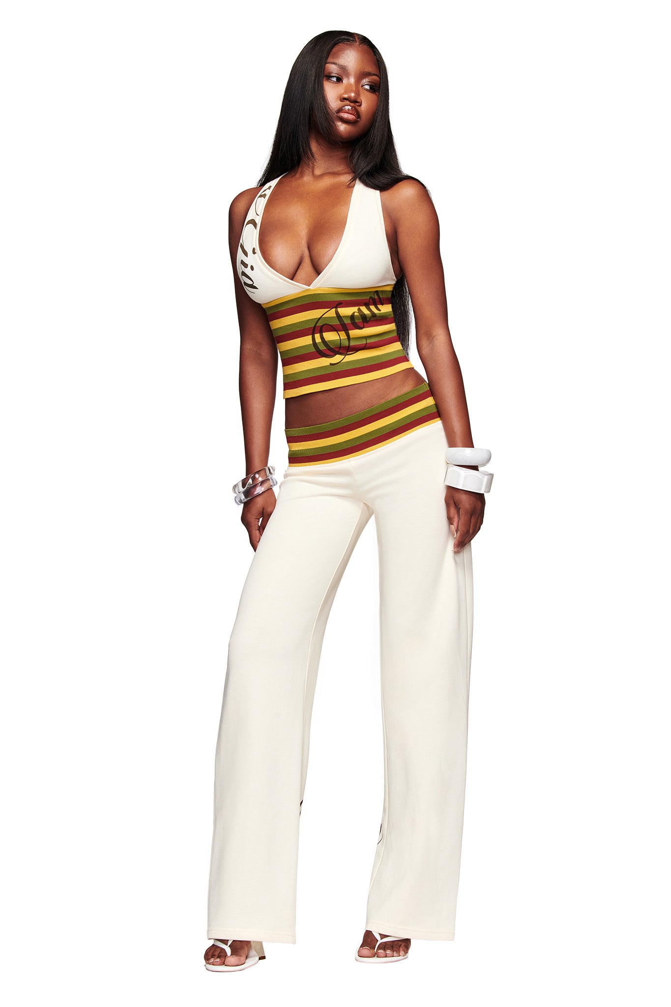 A woman poses confidently in the I.AM.GIA CANDI TOP - CREAM, a sleeveless V-neck racer back with “Oui” lettering, paired with wide-legged white pants and bold white bangles, her long straight hair contrasting the white background.