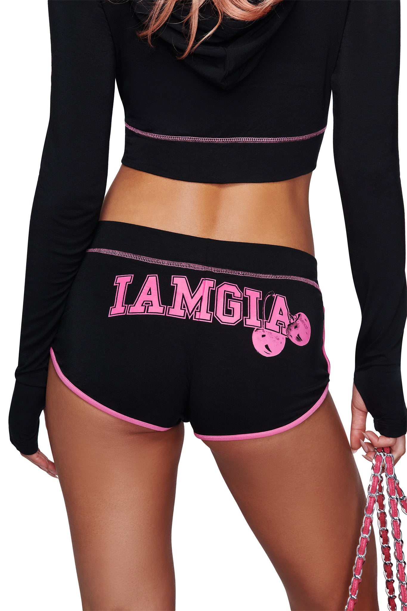 A person stands with their back to the camera, holding a pink chain in one hand, wearing the I.AM.GIA BLARE DOLPHIN SHORT in black with pink trim and "IAMGIA" printed on the back.
