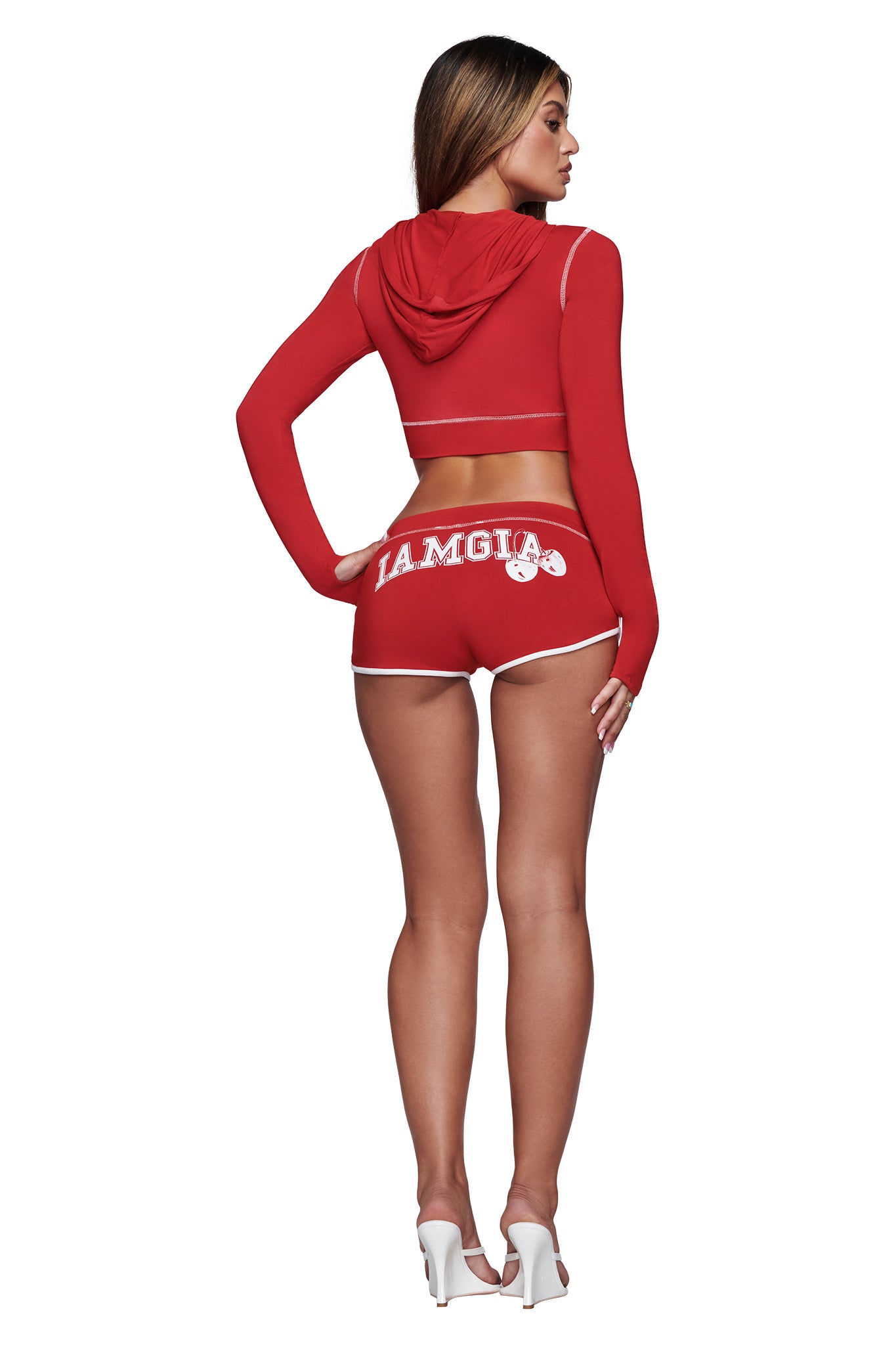 A woman is seen from behind, wearing the I.AM.GIA BLARE CROPPED HOODIE in red with matching shorts featuring the "I.AM.GIA" logo on the back, styled with white high heels.
