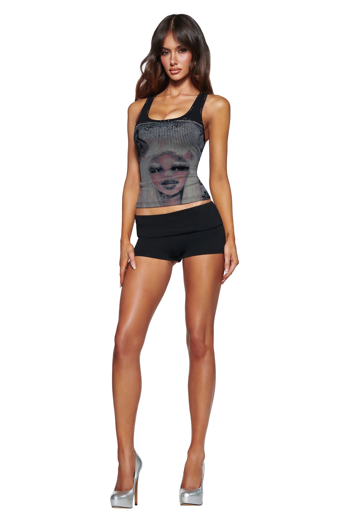 A woman with long brown hair wears the I.AM.GIA T-REX INA TANK - BLACK and black shorts, standing against a white background in silver high heels.