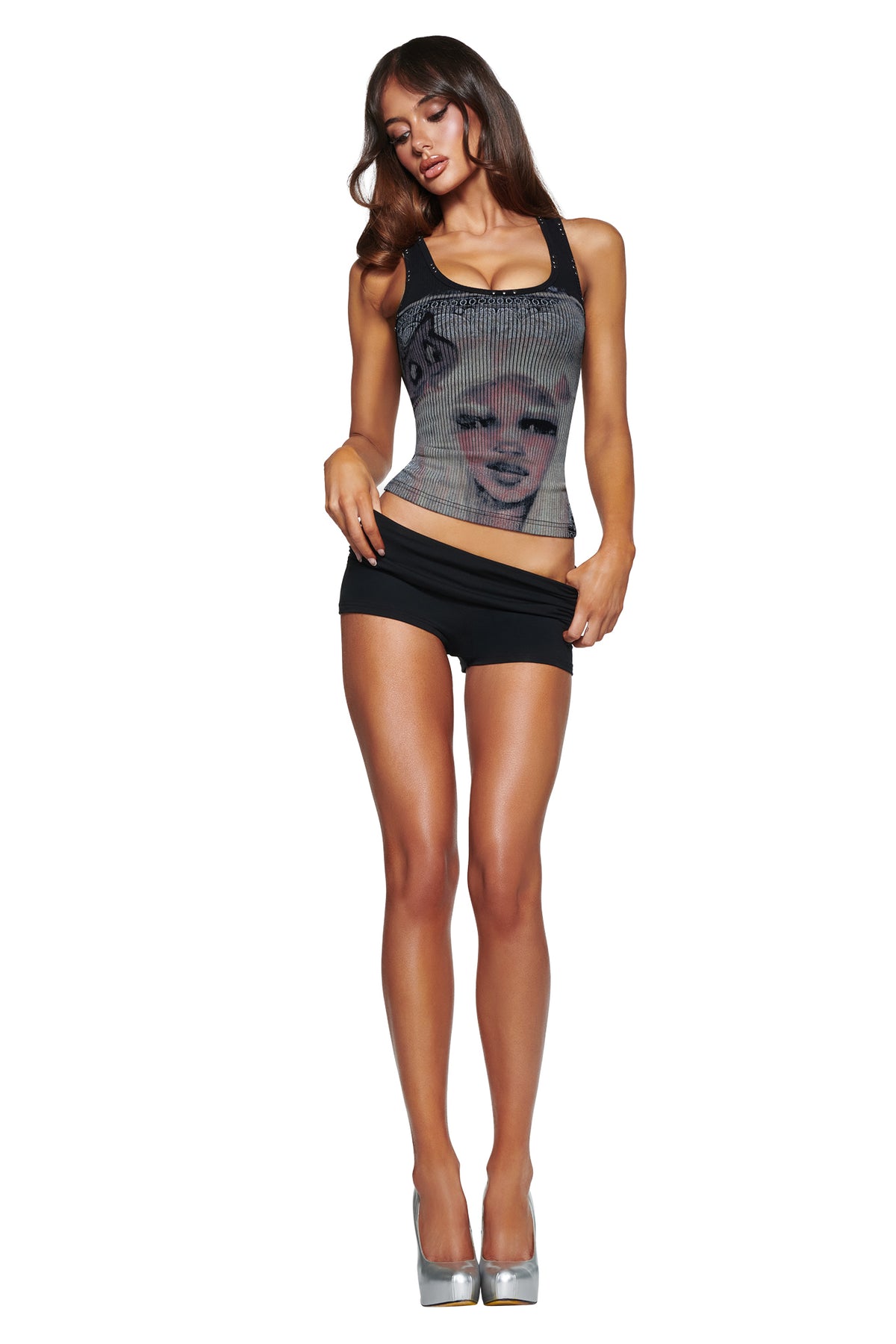 A woman with long brown hair wears the T-REX INA TANK - BLACK by I.AM.GIA, paired with black shorts and silver high heels, standing against a white background.
