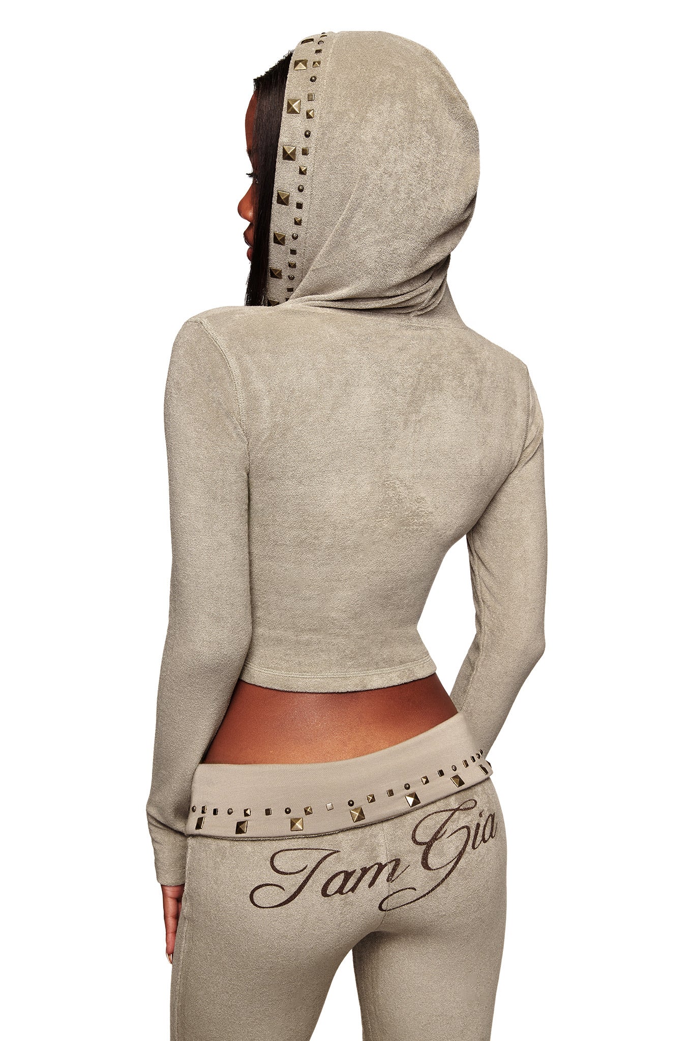A woman faces away from the camera, wearing the I.AM.GIA BLARE STUDDED TERRY HOODIE in stone and matching pants with "I am Gia" on the back waistband.