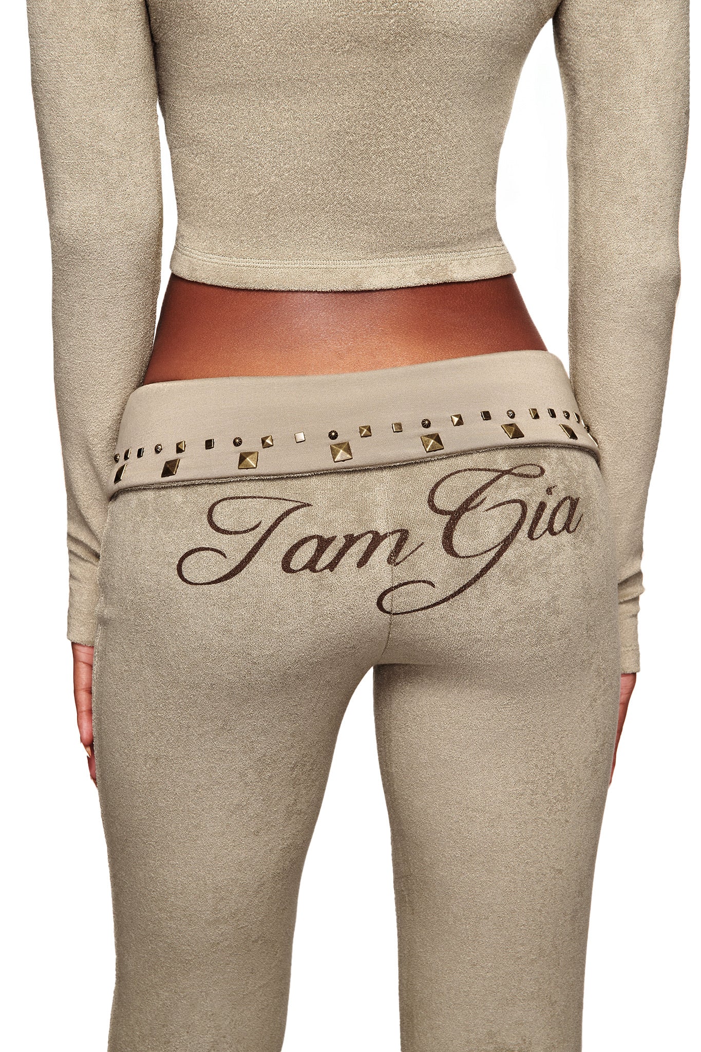 A person wears a beige, long-sleeve crop top with the I.AM.GIA BLARE STUDDED TERRY PANT in Stone, featuring a script logo on the back and studded belt detail at the waist. The photo is taken from behind.