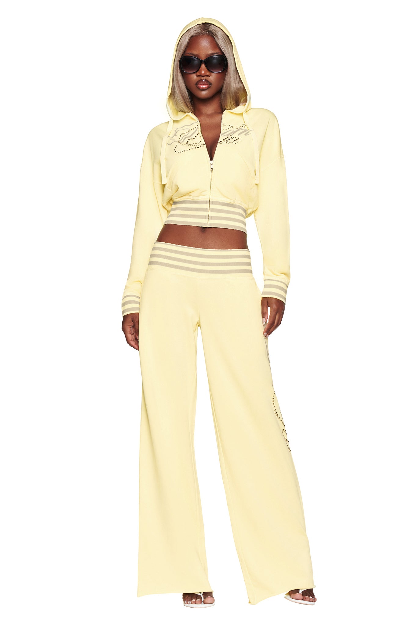 A woman poses confidently against a white background wearing the I.AM.GIA VEDA JACKET in yellow—a pale yellow cotton-blend fleece zip-up hoodie—paired with flared pants, sunglasses, and heeled sandals.