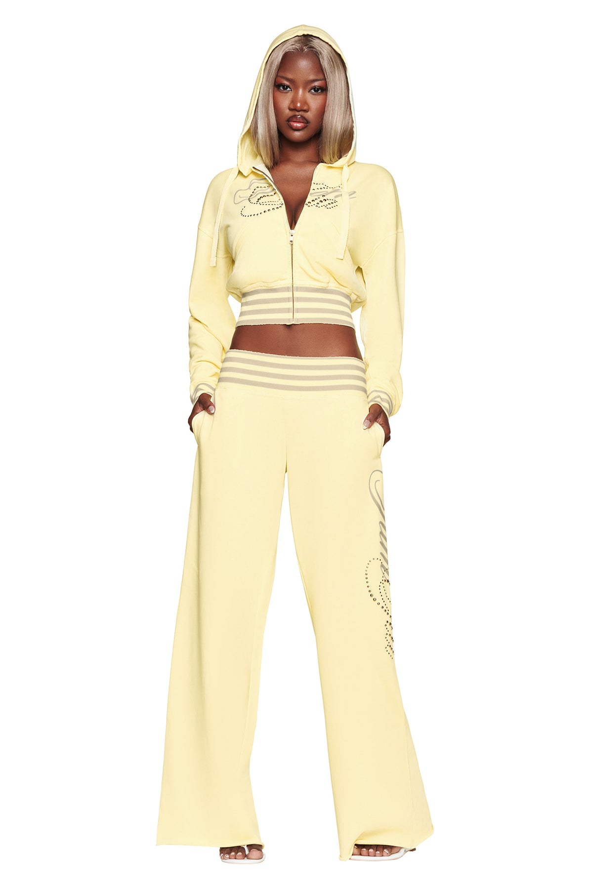 A woman wearing the I.AM.GIA VEDA JACKET in yellow—a light yellow cotton-blend fleece zip-up crop jacket—styled with matching wide-leg pants featuring embellishments, stands with her hands in her pockets against a white background.
