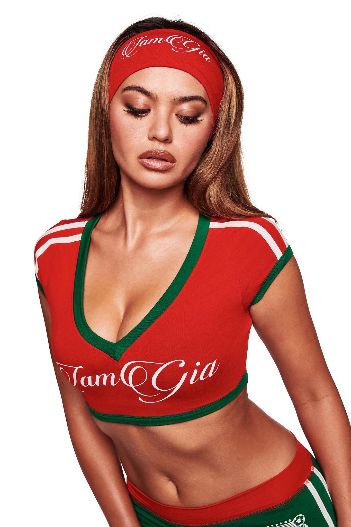 A woman with long brown hair wears the I.AM.GIA BLARE TWO-PIECE SPORT CROP TEE in crimson, pairing it with a matching red headband. The crop top features sporty green and white accents and prominent "I.AM.GIA" lettering. She looks down neutrally.