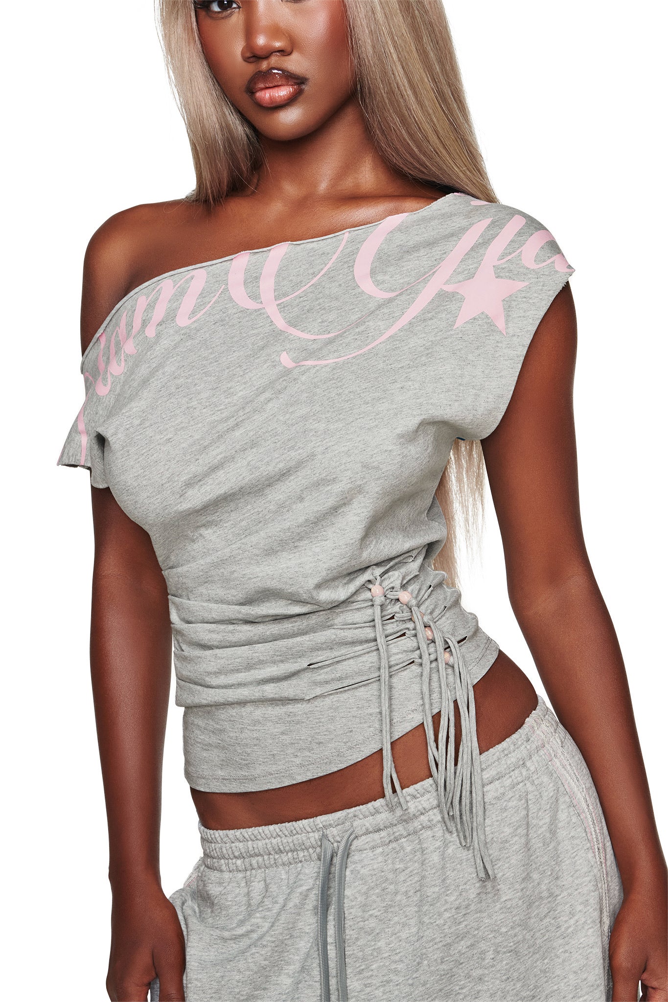 A woman wears the I.AM.GIA BEX Off Shoulder Top in grey, featuring pink lettering and drawstring sides, paired with matching gray sweatpants.
