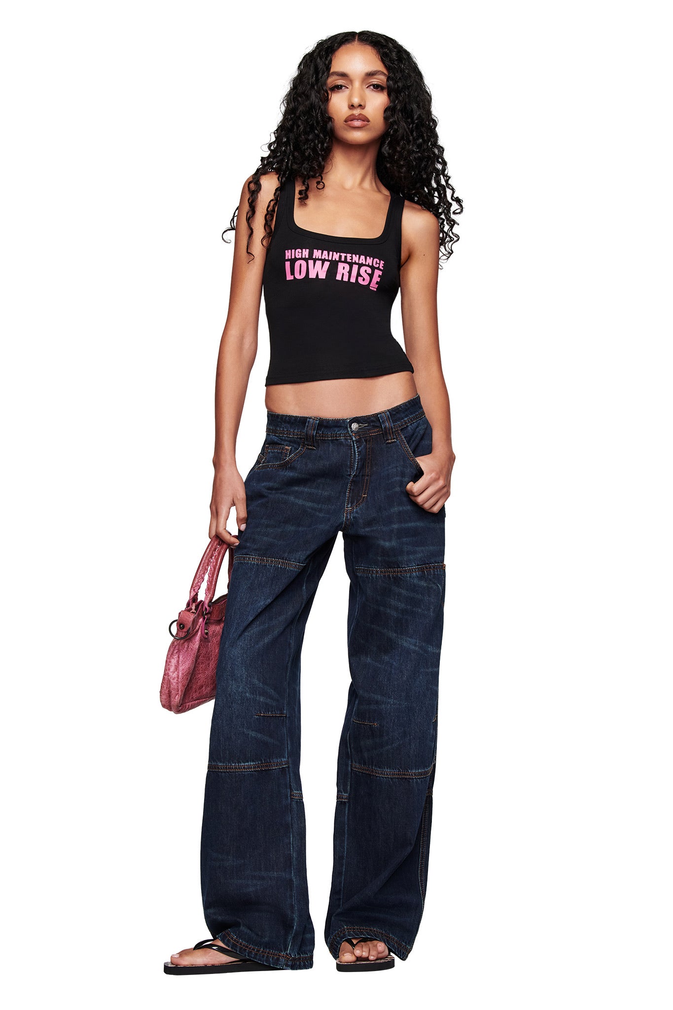 A woman with long curly hair stands in a relaxed pose, wearing I.AM.GIA’s MISTY JEAN in BLUE DUSTY WASH, a black tank top with "HIGH MAINTENANCE LOW RISE," black flip-flops, and carrying a pink handbag.