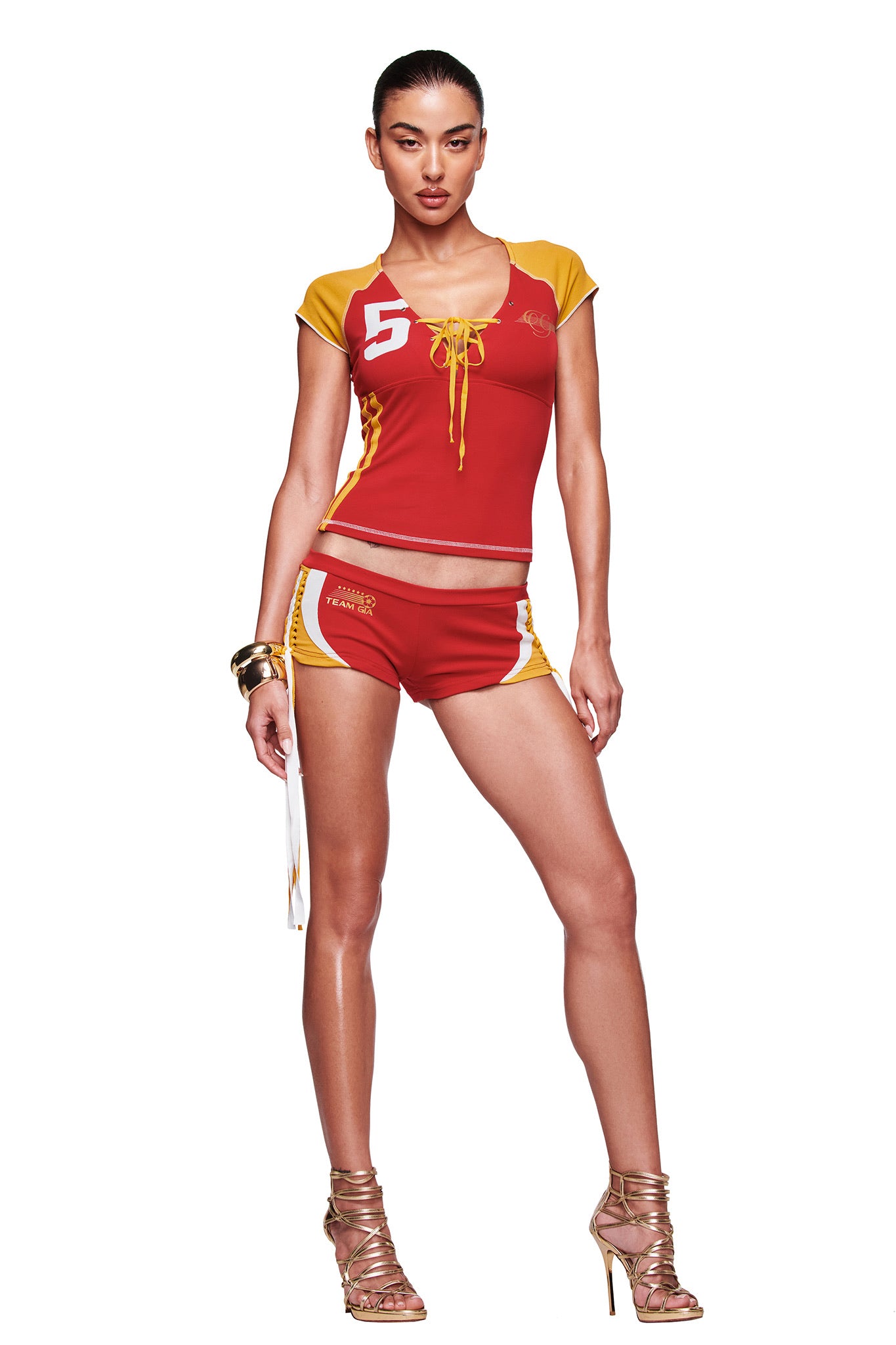 A woman poses confidently against a white background in I.AM.GIA’s NOVIA MICRO SPORT SHORT - RED, paired with a sporty red and yellow top featuring the number 5, gold high-heeled sandals, and gold bangles.