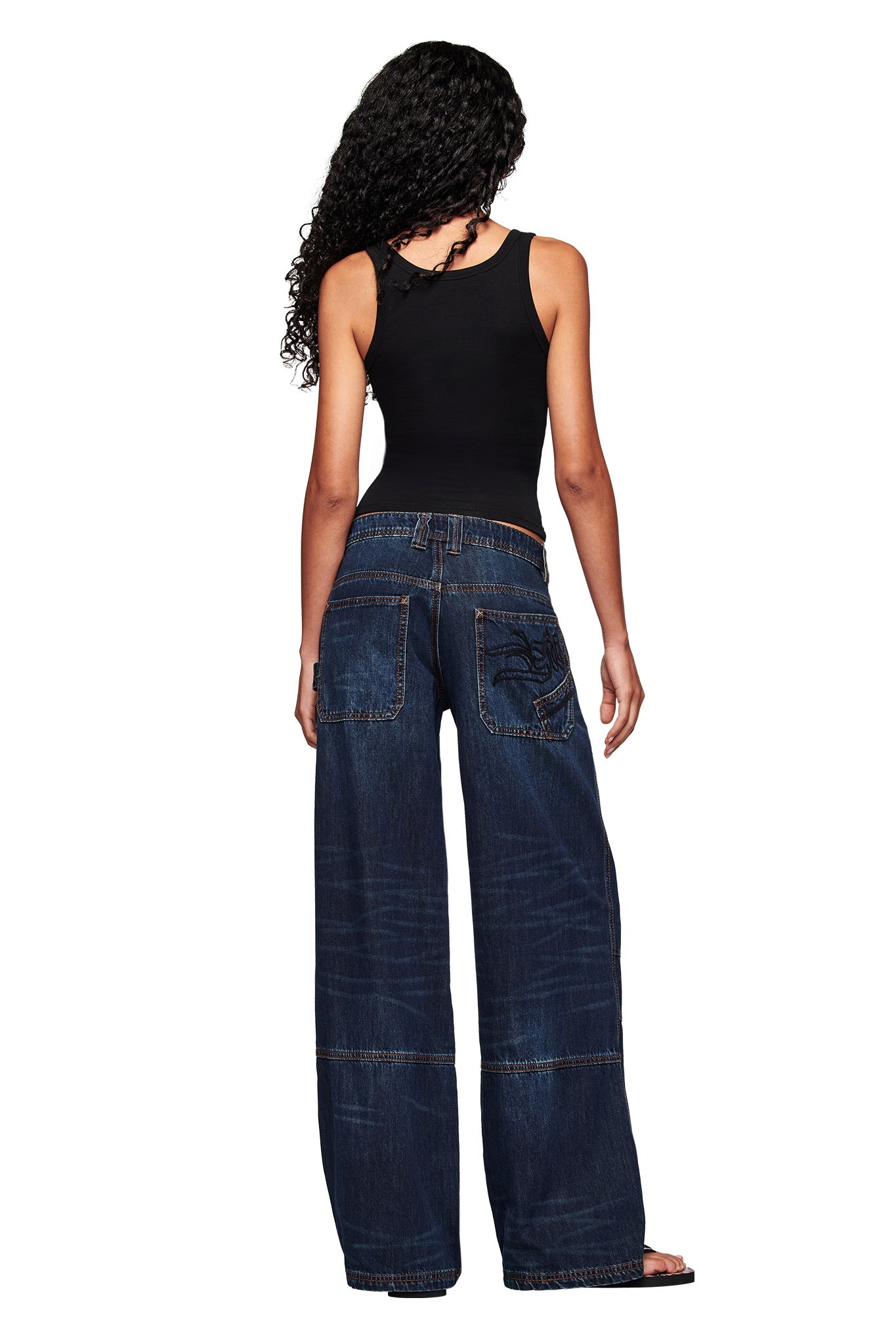 A woman with long curly hair, wearing a black sleeveless top and I.AM.GIA MISTY JEAN in blue dusty wash, stands facing away from the camera against a white background.