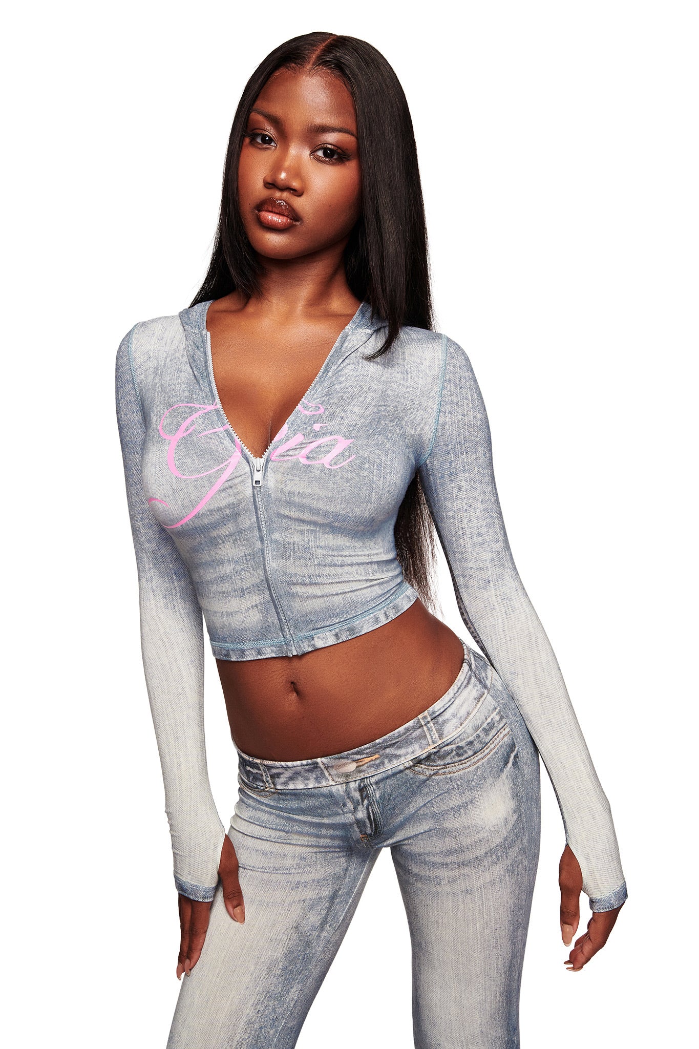 A woman with long straight hair wears the BLARE HOODIE - DENIM TROMPE L'OEIL by I.AM.GIA, featuring a zip front and pink "Gira" script, paired with matching jeans. She poses with one hand on her hip against a white background.