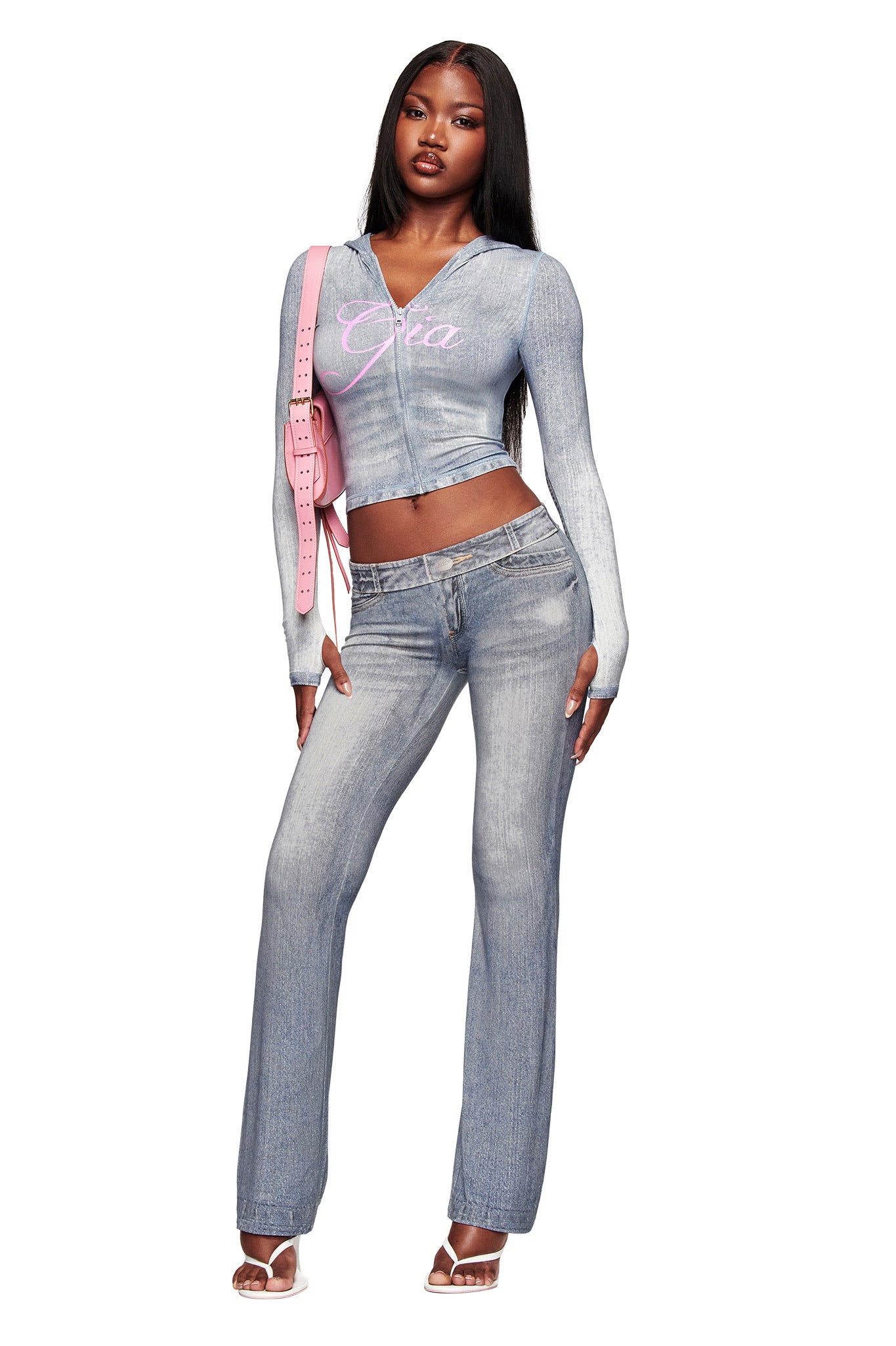A woman wears I.AM.GIA's BLARE TRACKPANT - DENIM TROMPE L'OEIL, paired with a matching cropped hoodie featuring "Gia" in pink, clear-heeled sandals, and a pink shoulder bag.