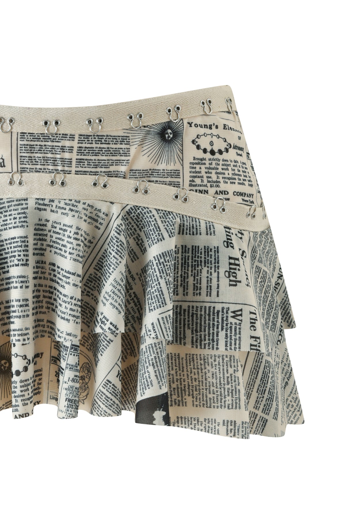 The SORA MINI SKIRT - LEFT ON READ by I.AM.GIA is a pleated mini skirt crafted from newspaper print fabric with black-and-white article designs and finished with metal grommet details along the waistband.