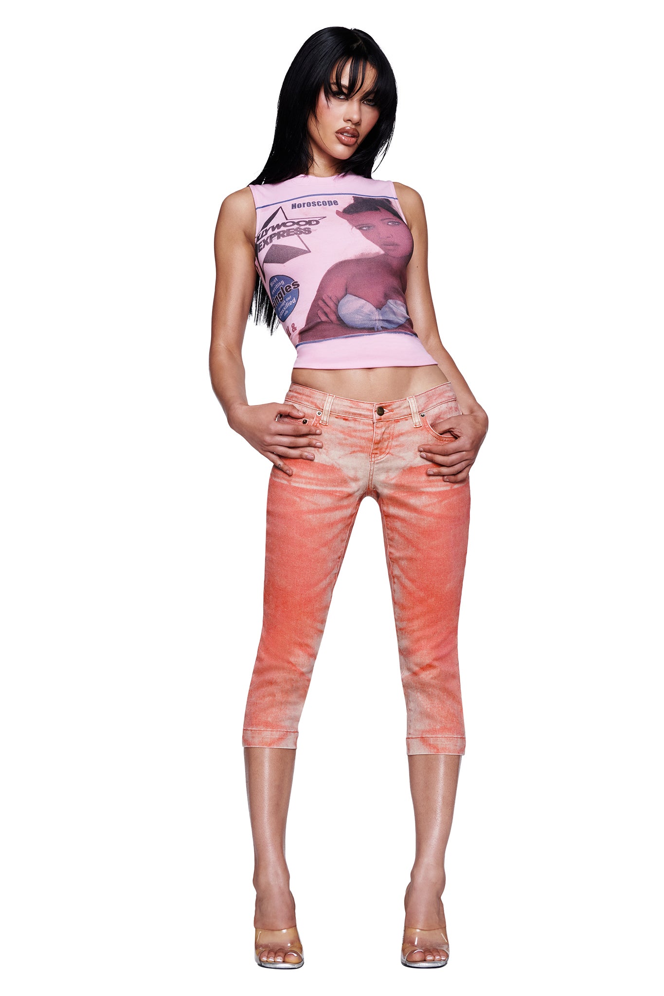 A woman wearing the I.AM.GIA SUNSET CAPRI JEAN - PEACH, paired with a pink graphic crop top and clear high-heeled shoes, stands with her hands in her pockets against a white background.